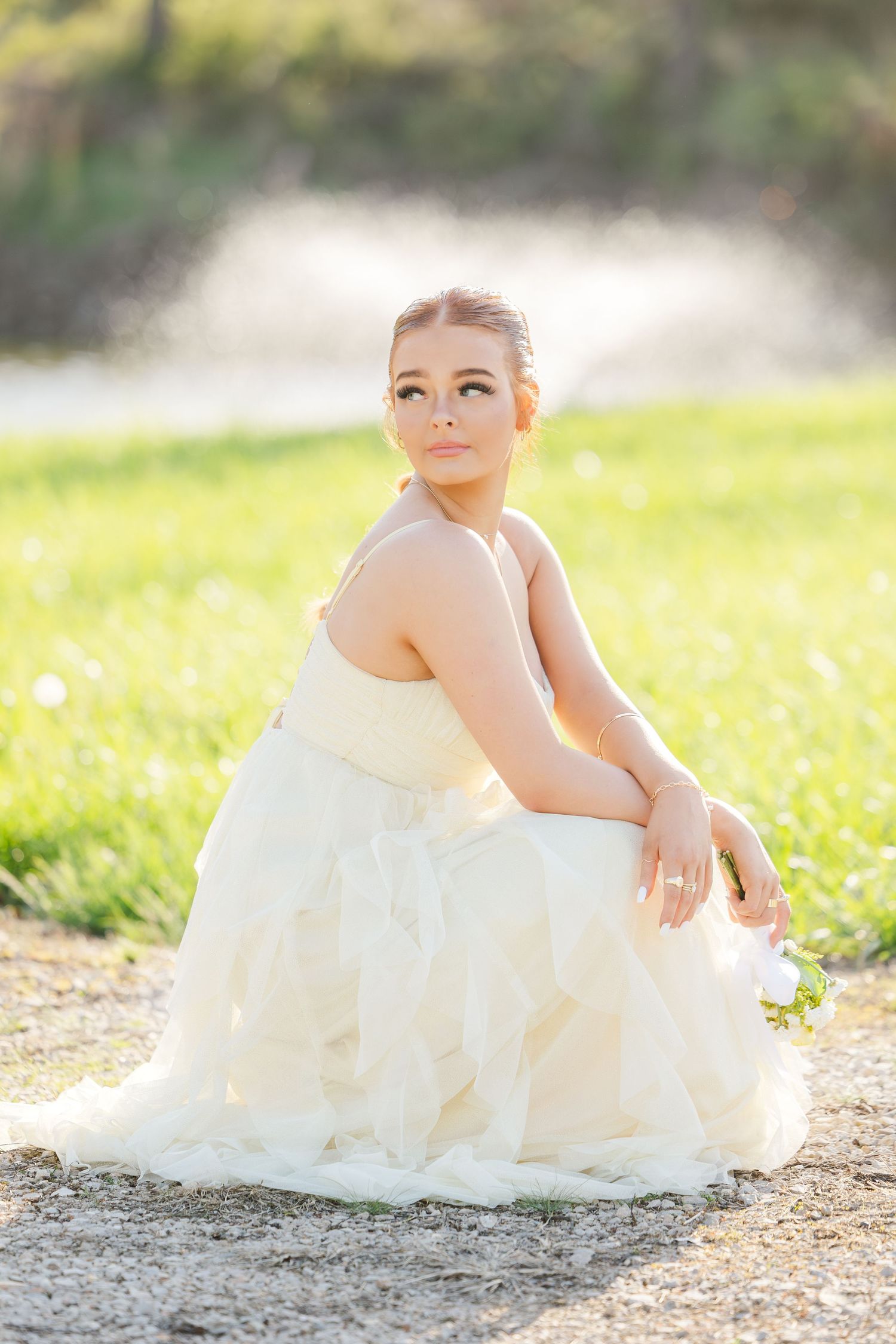 A person in a flowing white gown sits gracefully on the ground in a sunlit outdoor setting with grass and dandelions.