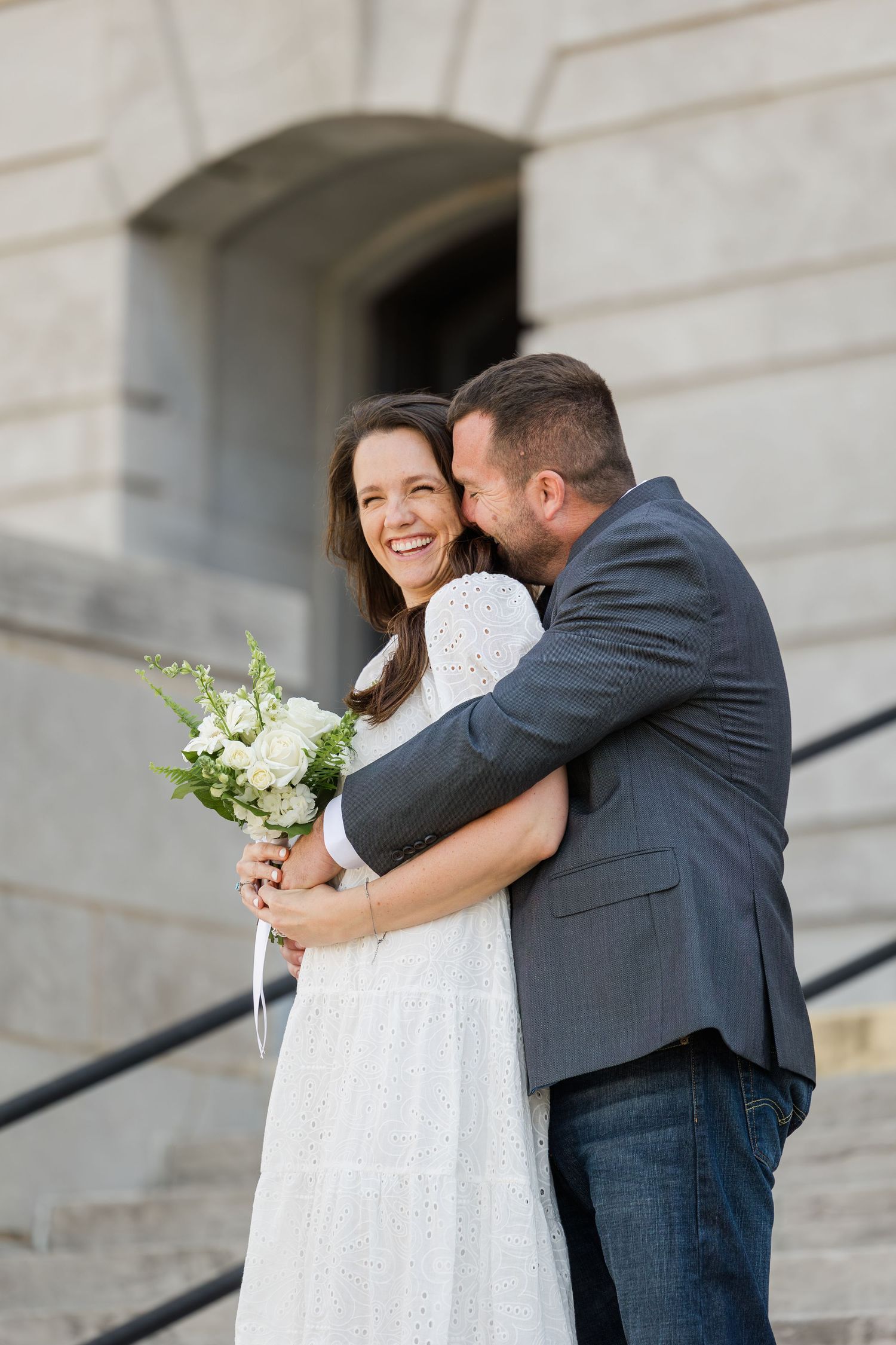 A joyful couple shares a tender moment on stone steps, with the bride wearing white and holding a delicate bouquet.