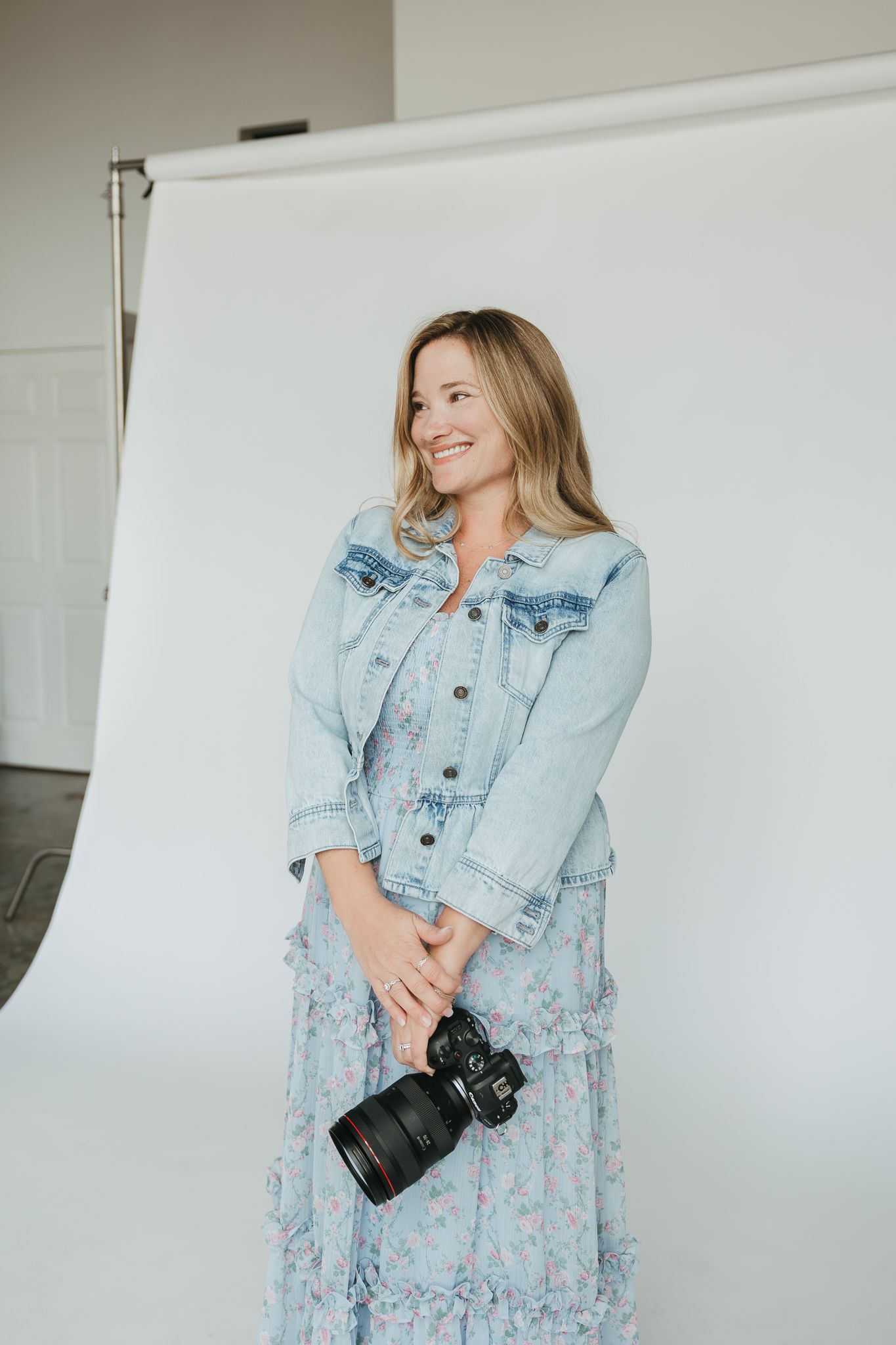 Fashion blogger poses in floral spring dress and light denim jacket while holding a professional camera in studio setting.