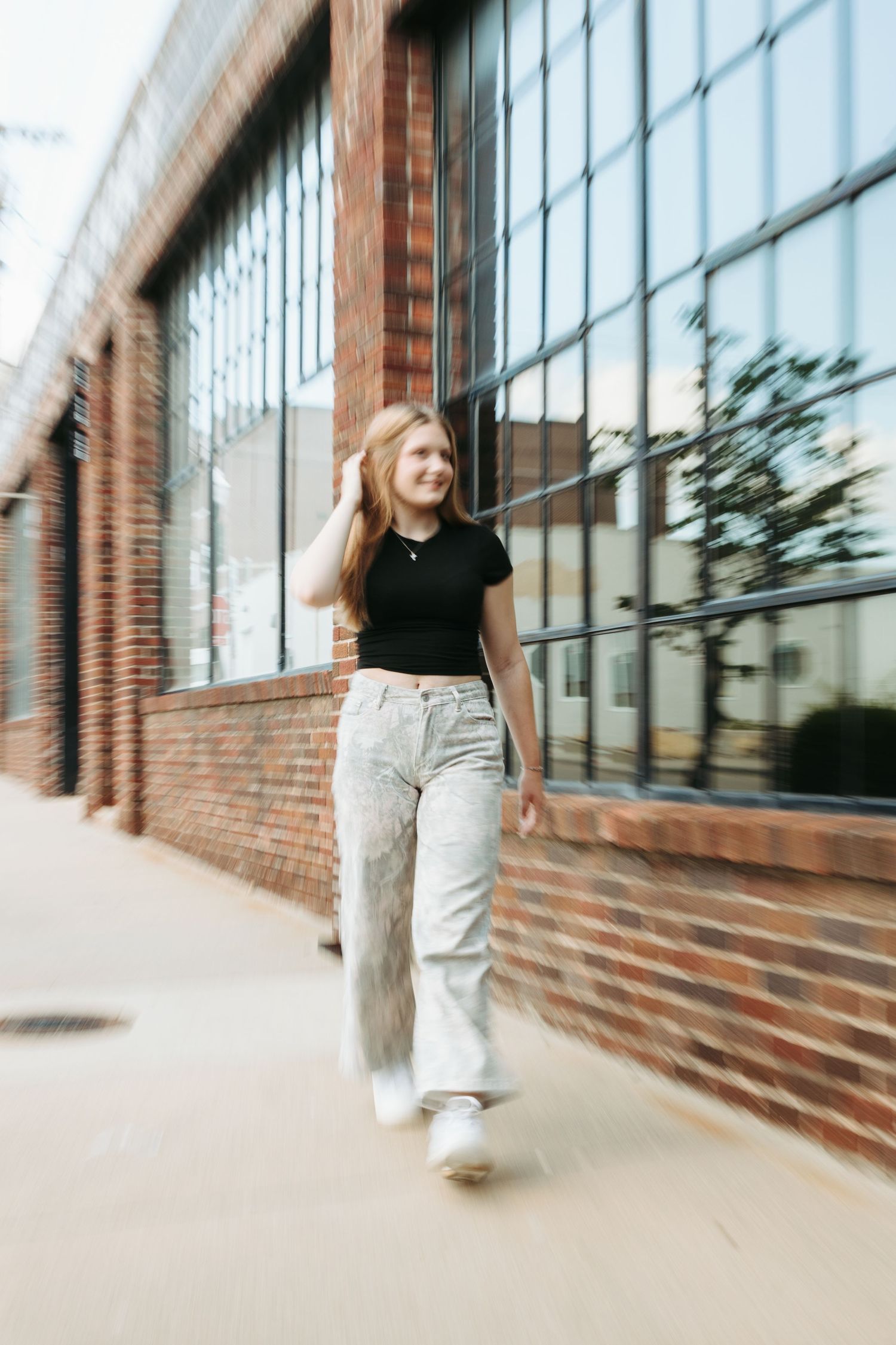 A casual street style photo showing someone in a black t-shirt and light jeans walking past brick and glass architecture.
