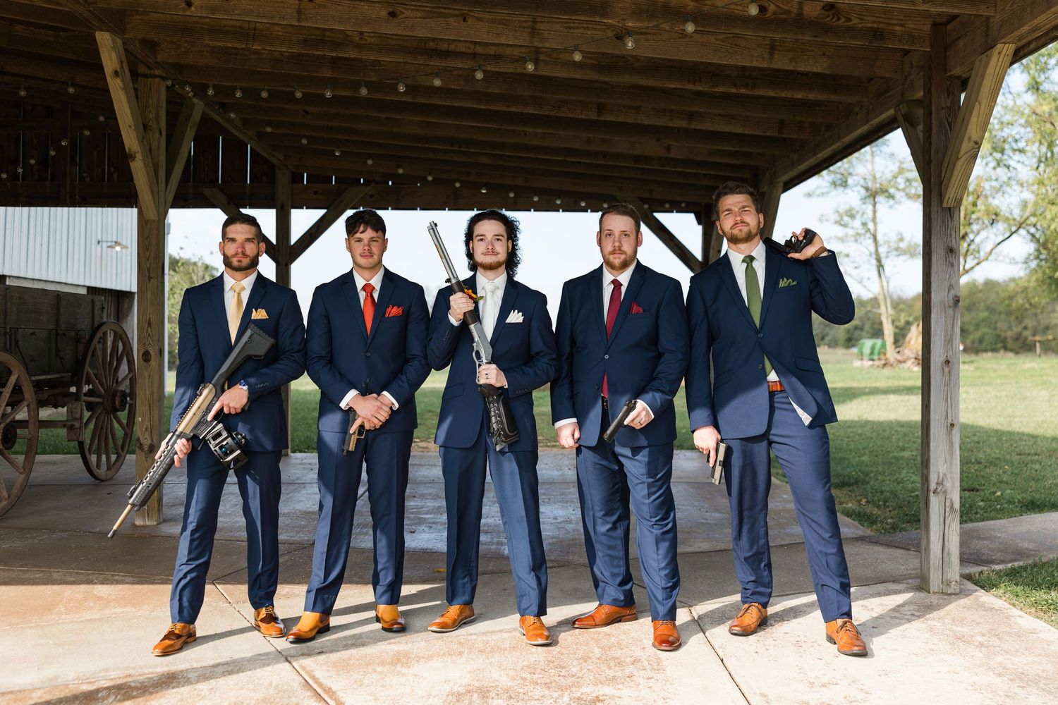 Groomsmen in navy suits show off colorful socks in creative wedding photo poses at rustic venue.