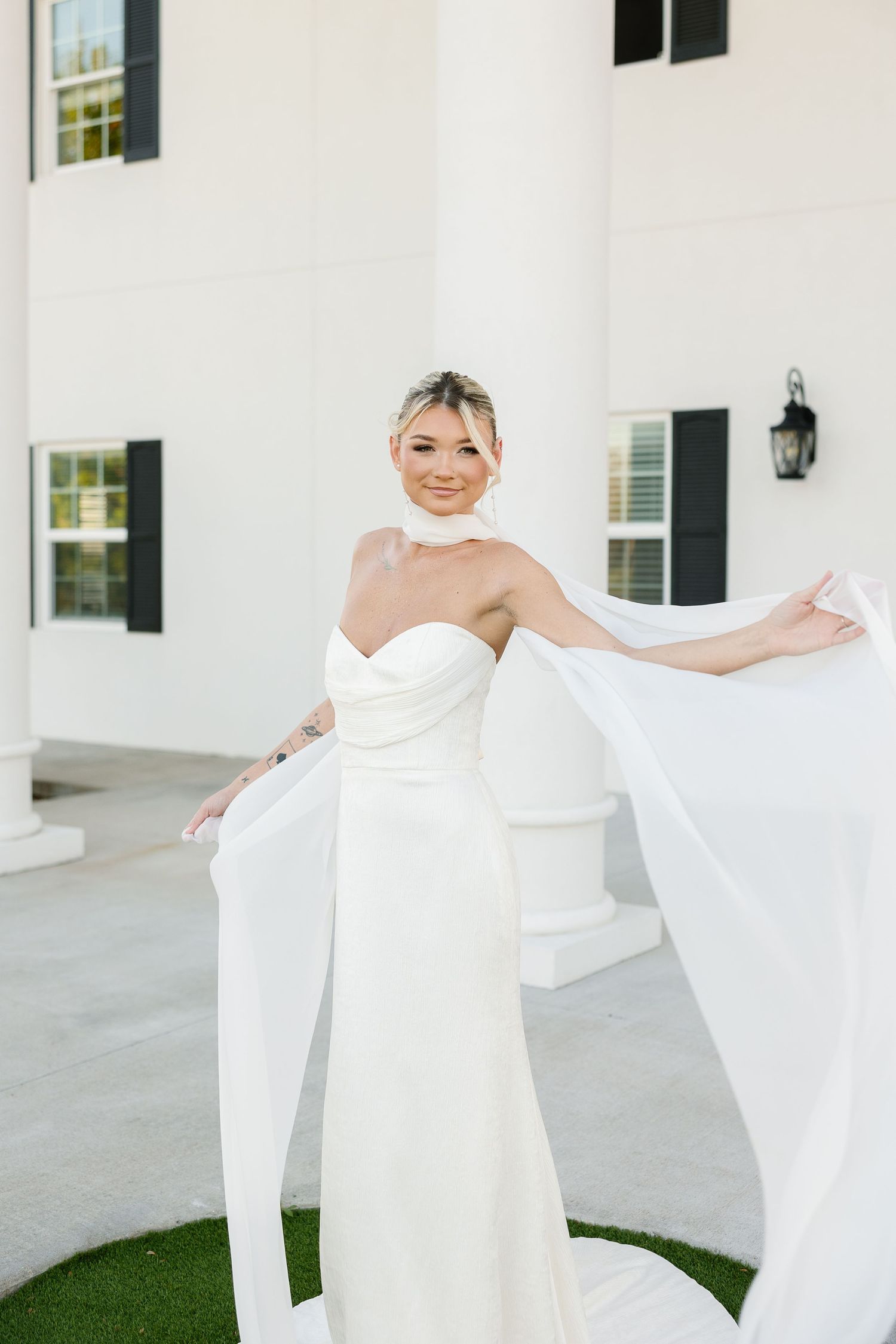 A person in an elegant white strapless wedding dress stands gracefully on a white porch, playfully holding the dress's flowing fabric.