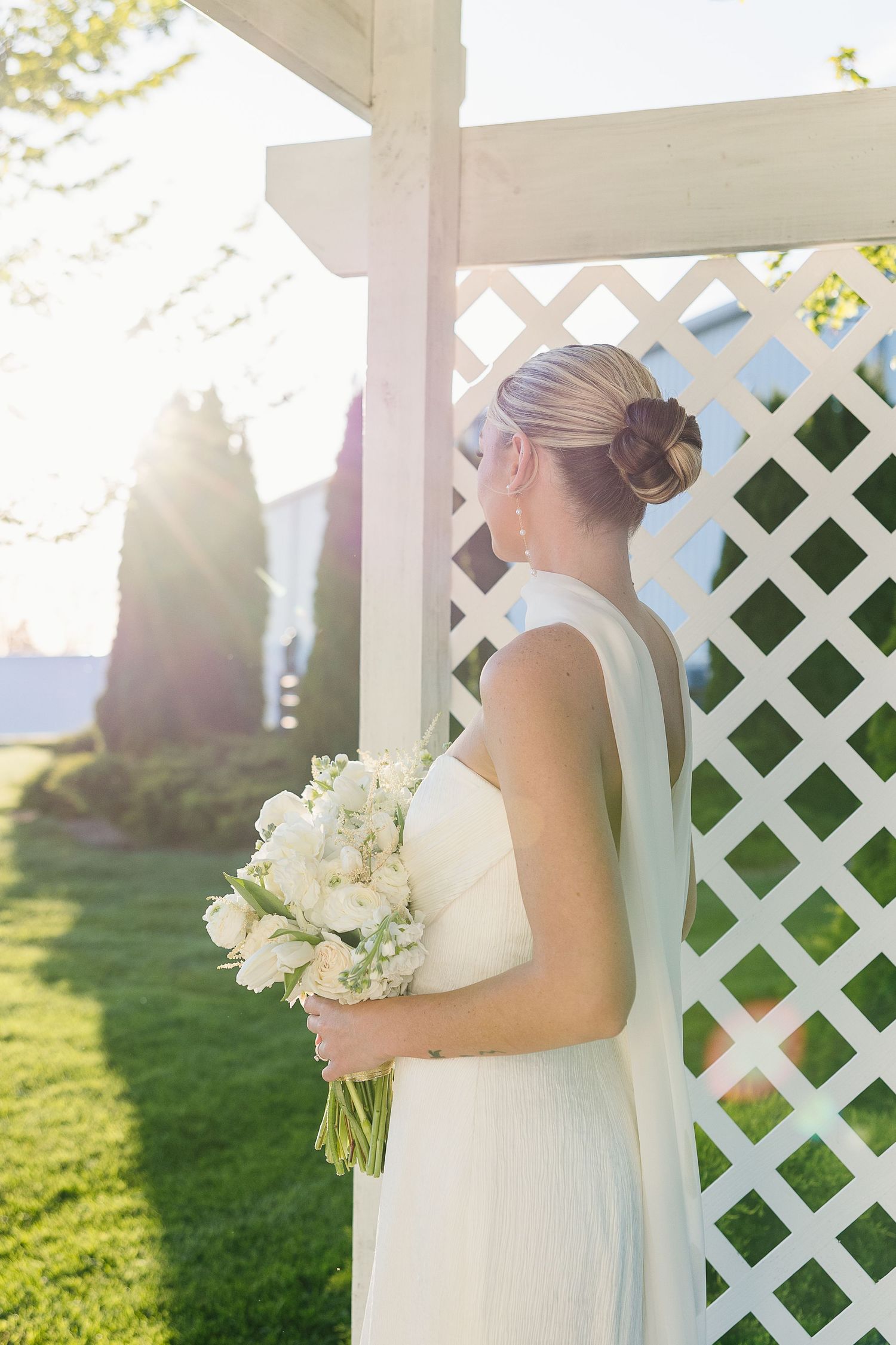 A person in an elegant white wedding dress holds a bouquet of white flowers beside a lattice pergola at sunset.