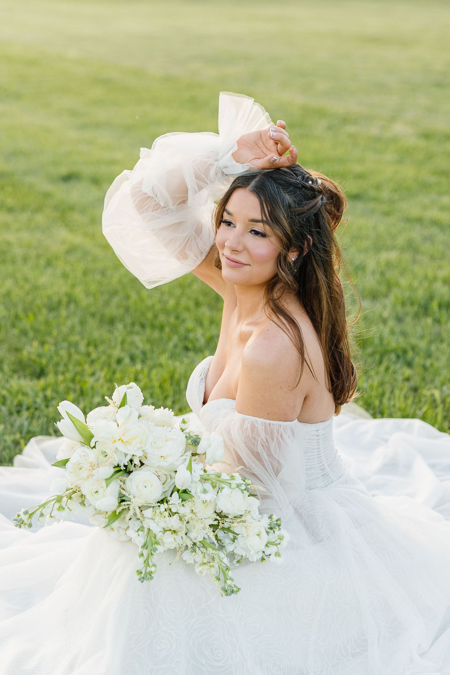 A person in a white wedding dress holds a delicate bouquet of white flowers while posing in a sunlit grassy field.