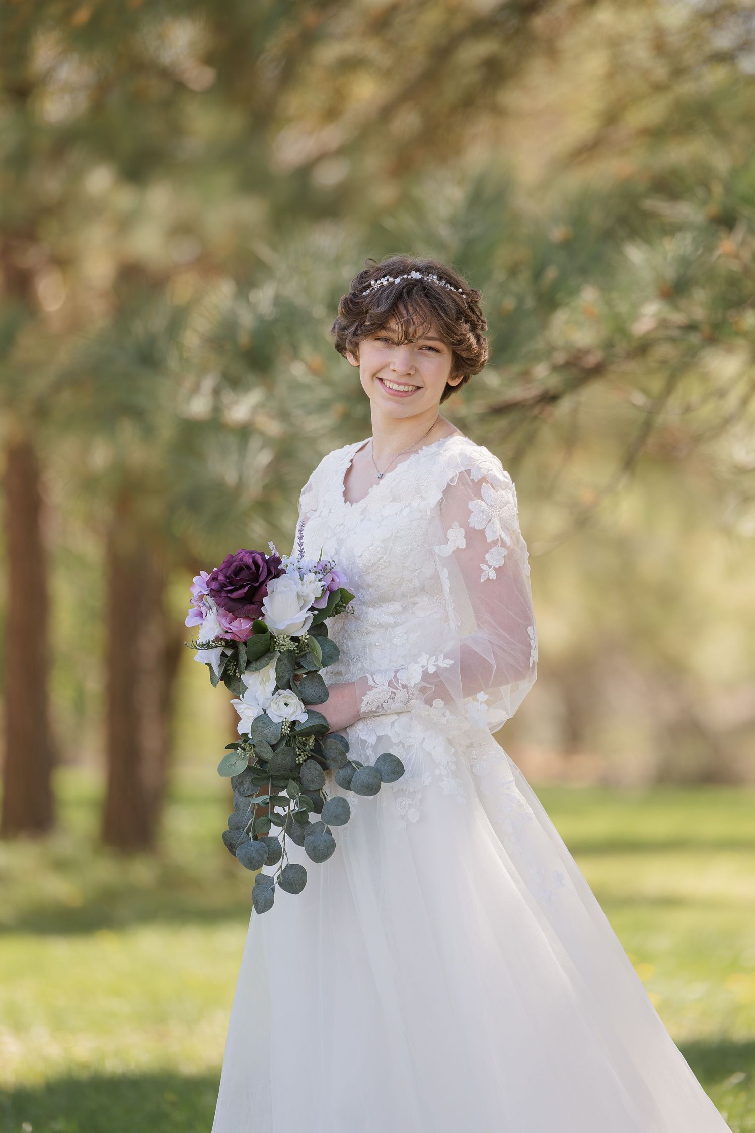A bride in a flowing white lace gown and veil holds a purple and white floral bouquet in an outdoor garden setting.