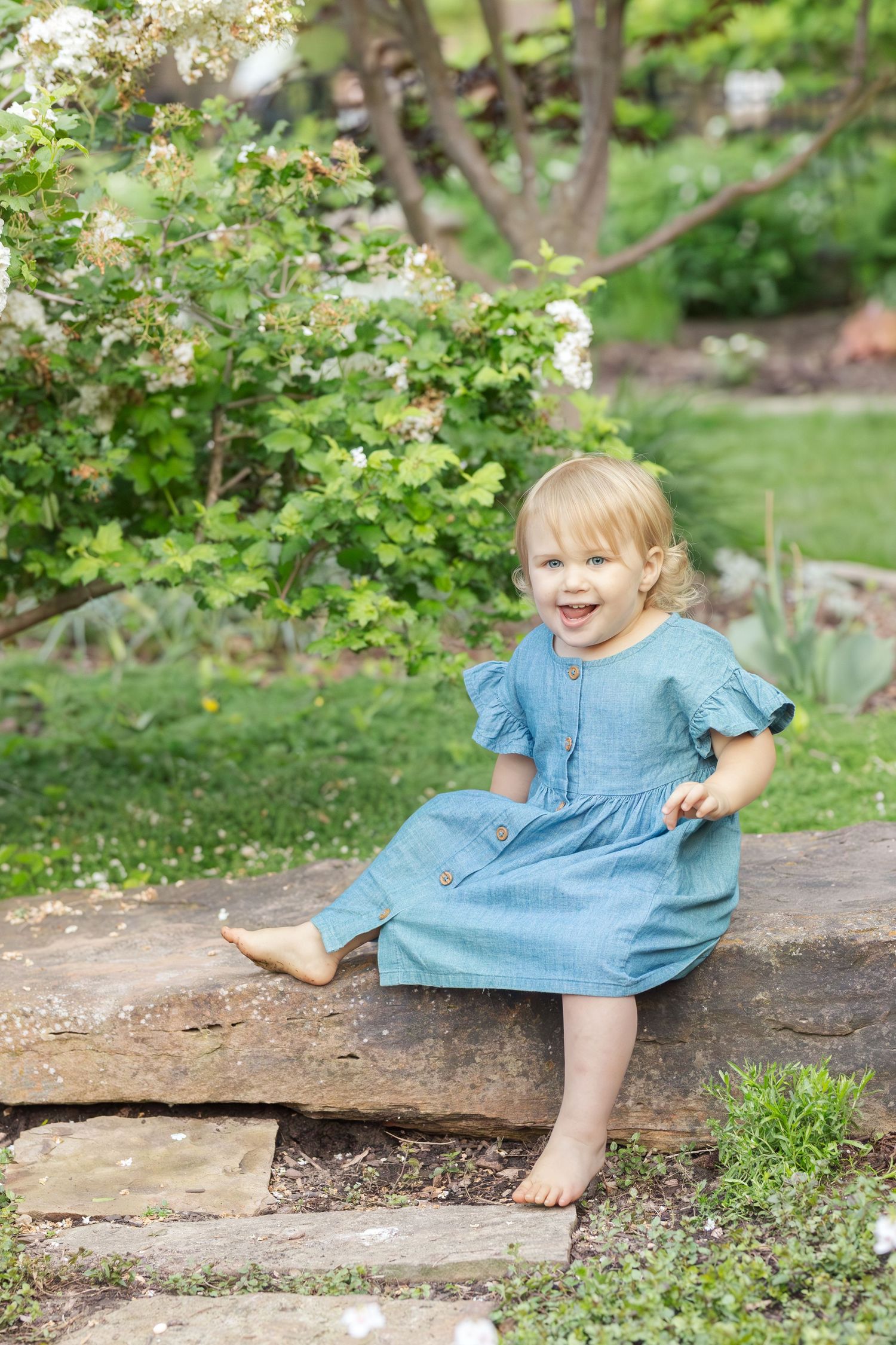 A joyful little one in a blue dress sits barefoot on stone steps in a lush garden setting.