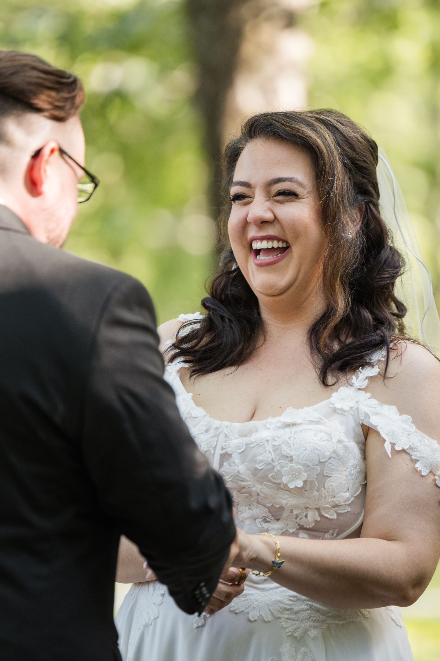 A joyful bride in a white lace dress laughs during an outdoor wedding ceremony.
