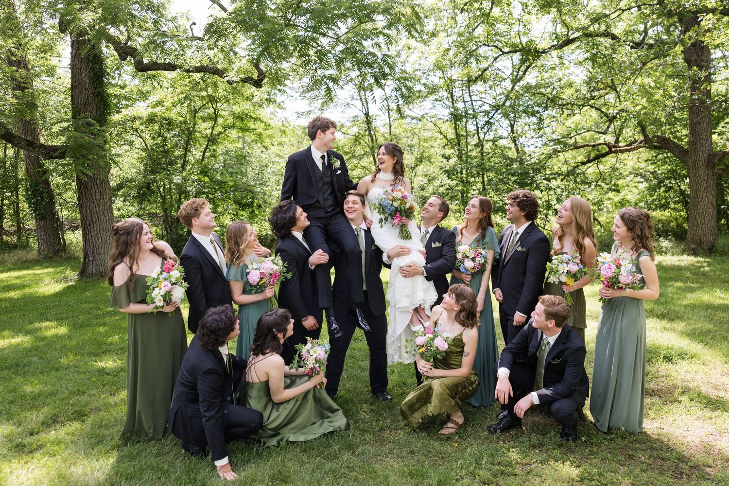 Wedding party in green dresses and black suits posing outdoors under trees on a sunny day.
