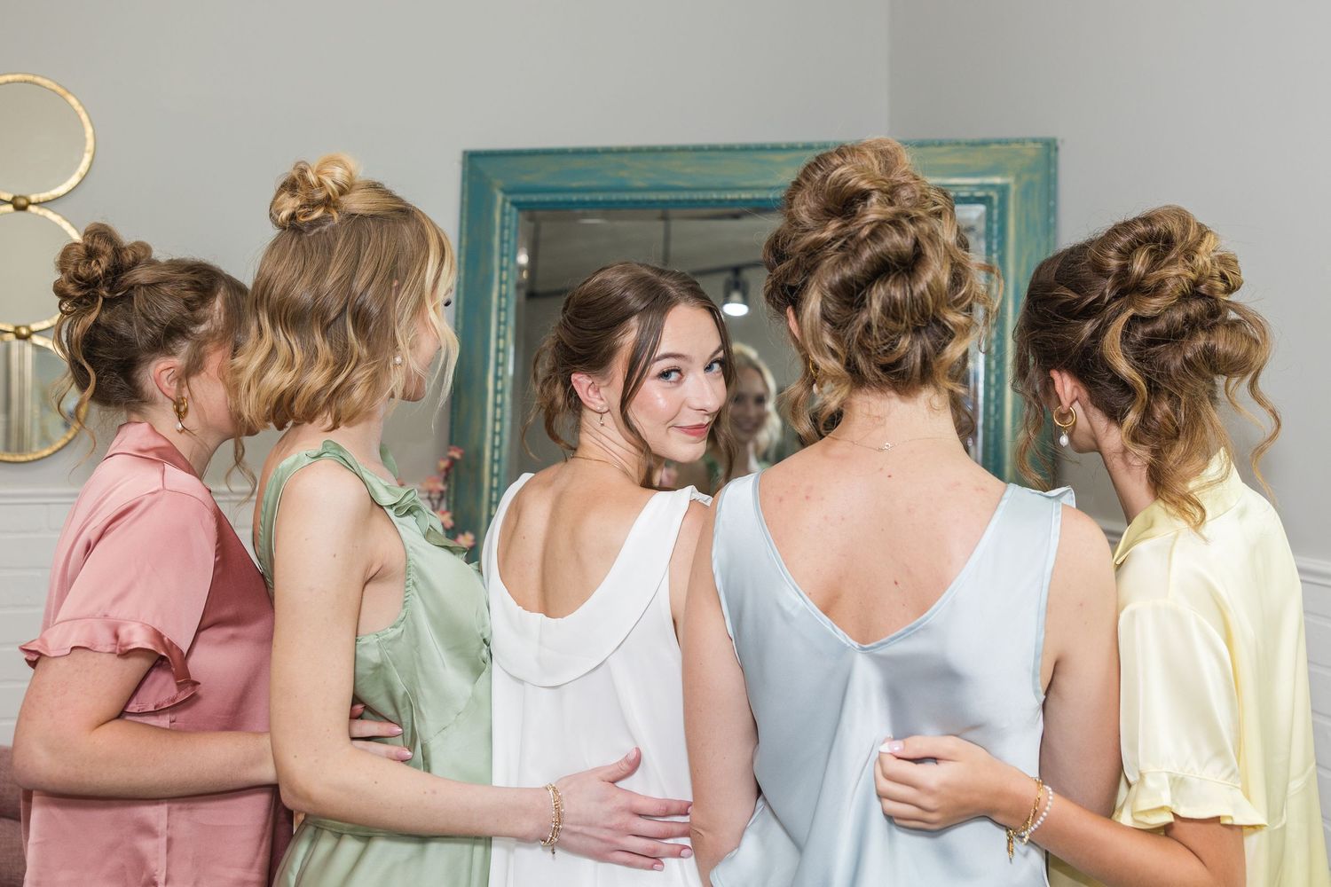 Bride surrounded by bridesmaids in pastel robes sharing a joyful getting-ready moment before the wedding.