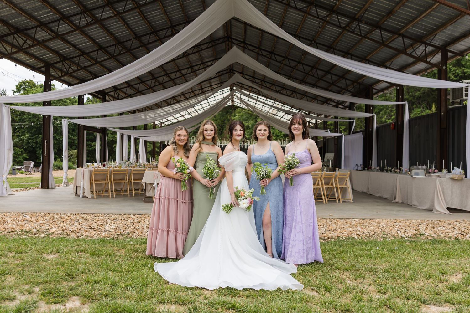 Bride in white gown poses with four bridesmaids in pink and lavender dresses under a draped outdoor wedding venue.