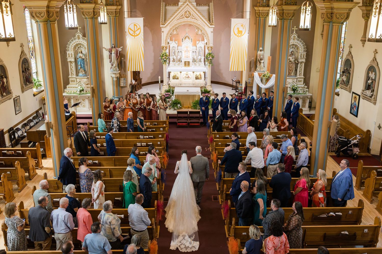 Bride walking down the aisle of a decorated Catholic church during a wedding ceremony with guests standing in pews.