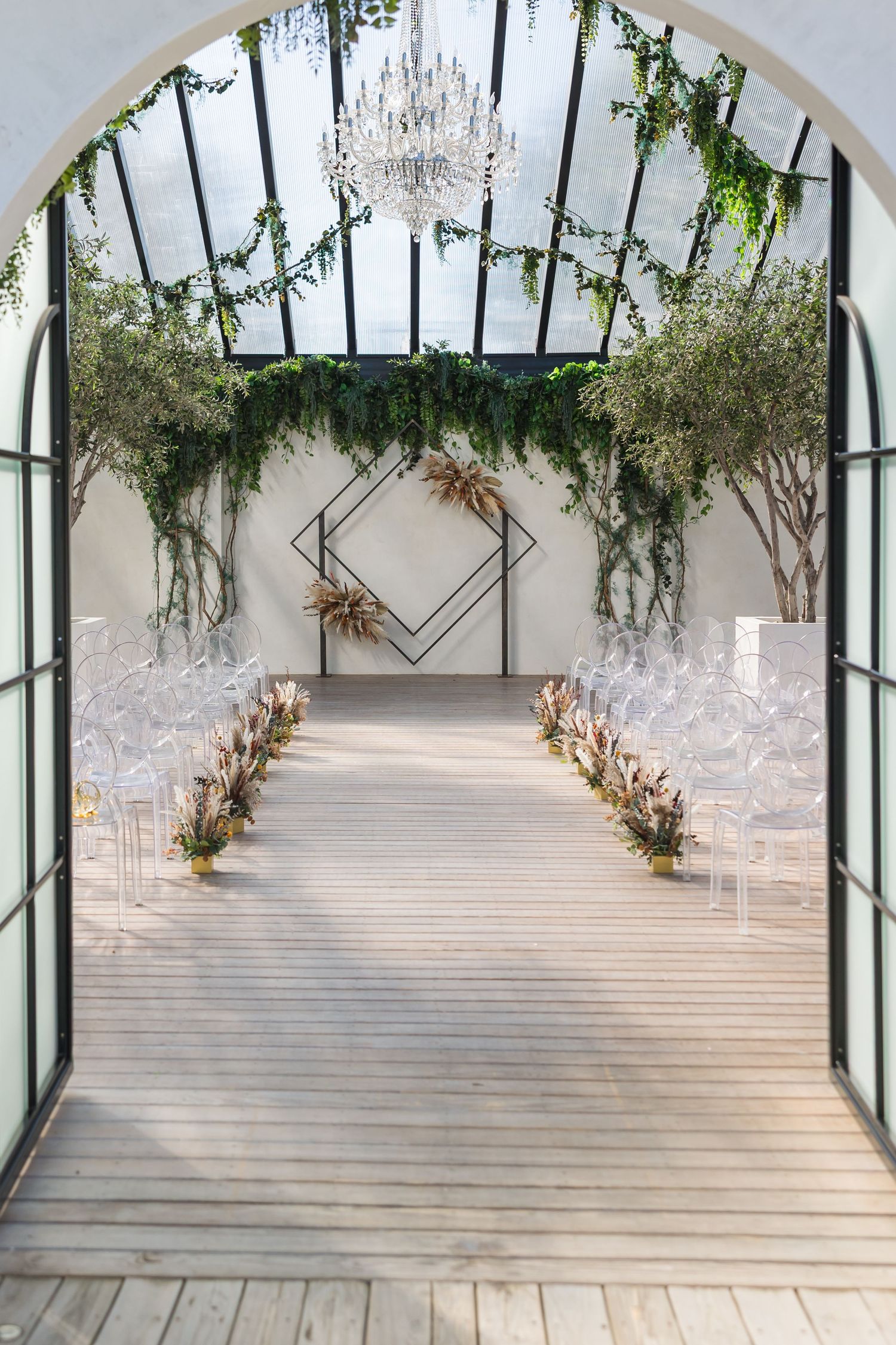 Elegant greenhouse wedding venue with chandelier, ghost chairs, floral aisle, and geometric arch backdrop.