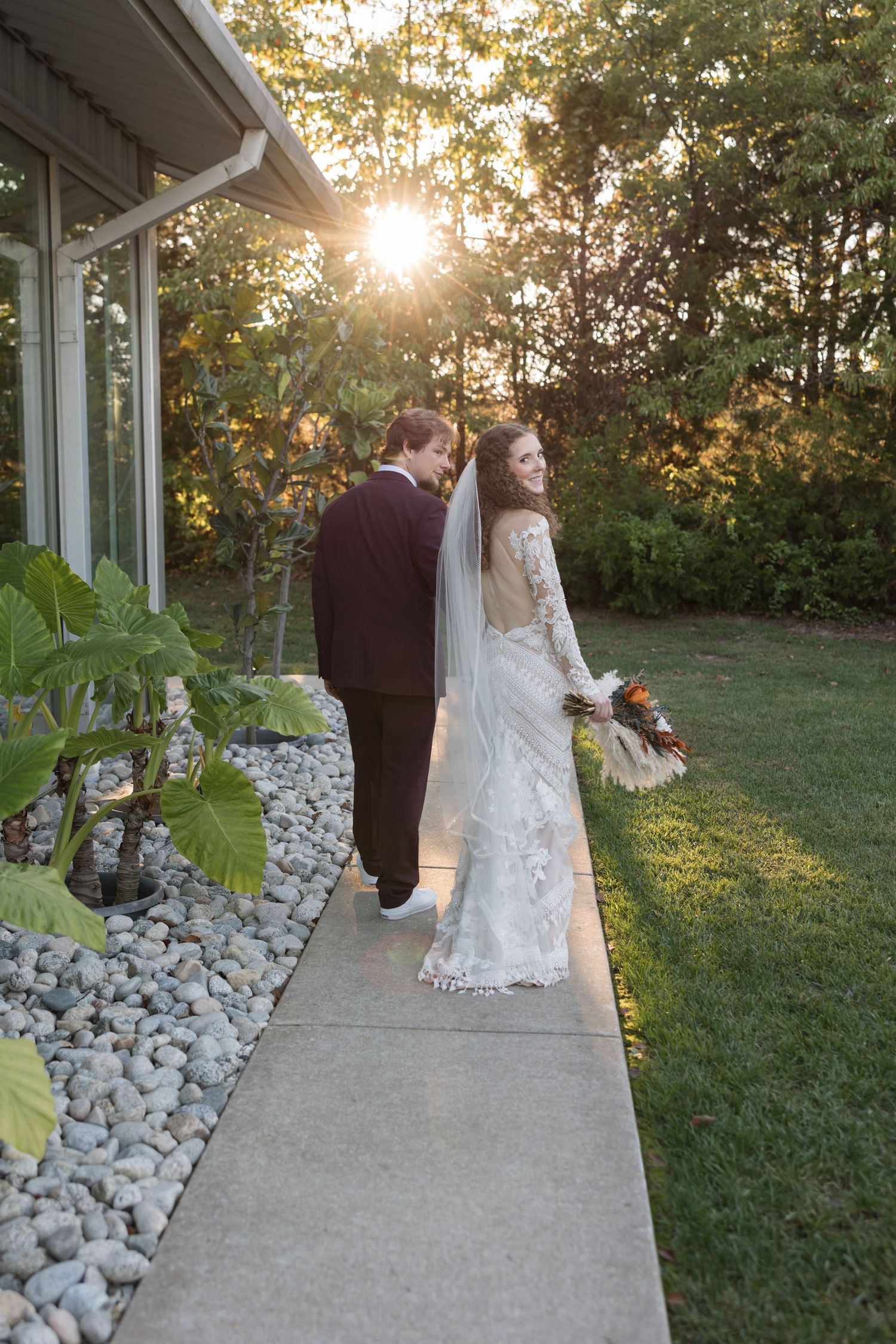 Bride in lace gown with veil and groom in burgundy suit walk at sunset along garden path.