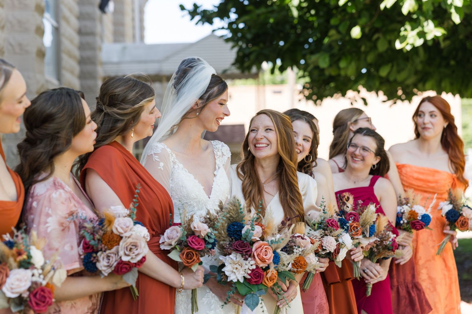 A bride in a lace gown and veil stands with bridesmaids in mismatched warm-toned dresses holding floral bouquets.