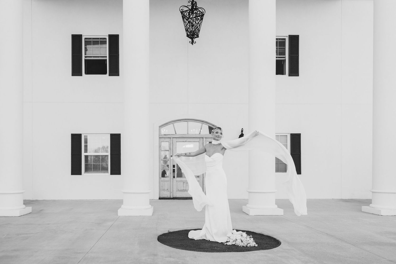 Bride in white gown twirling veil outside elegant white colonial venue with black shutters and grand columns.