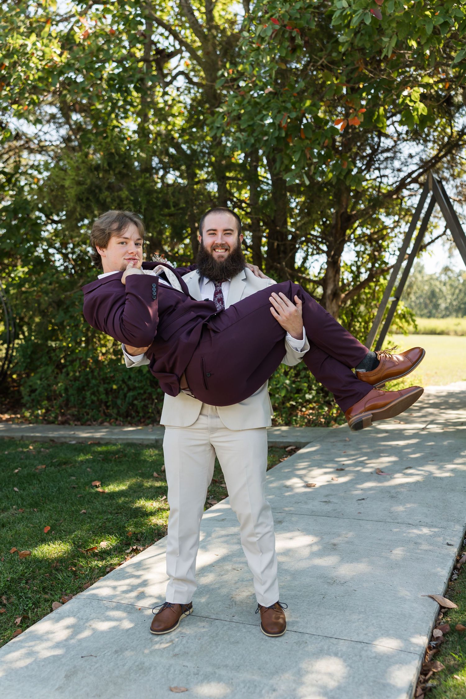 Bearded groomsman in cream suit carrying laughing man in burgundy suit outdoors at wedding.