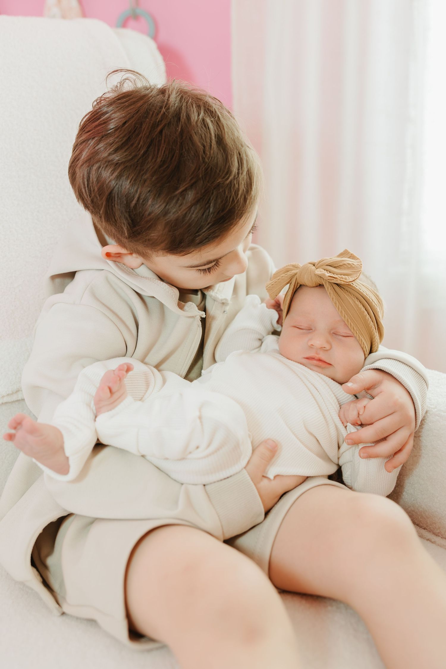 Two young children in cozy beige outfits share a tender moment together on a pink sofa.