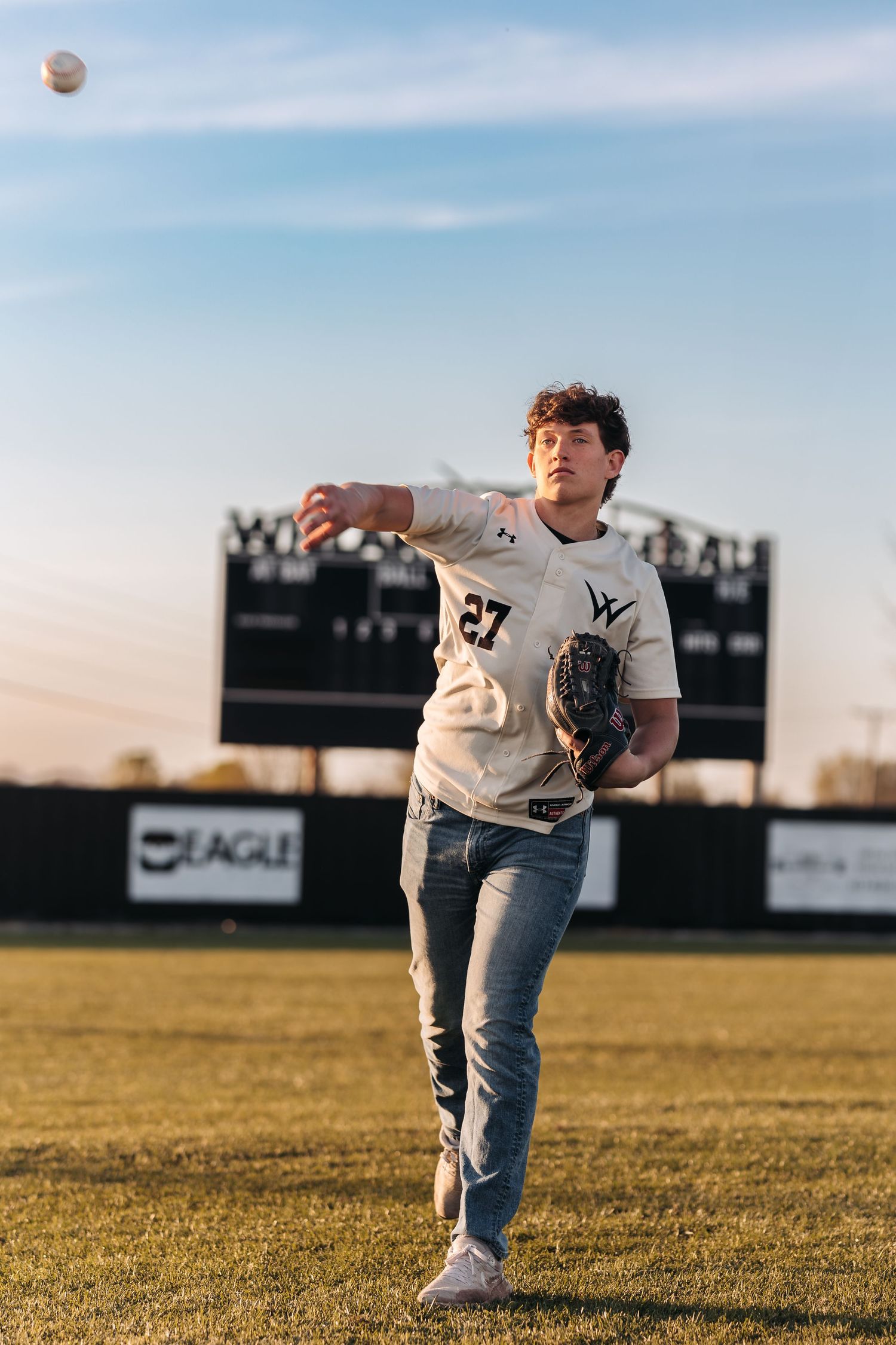Baseball player in white uniform throwing ball on field with scoreboard in background.