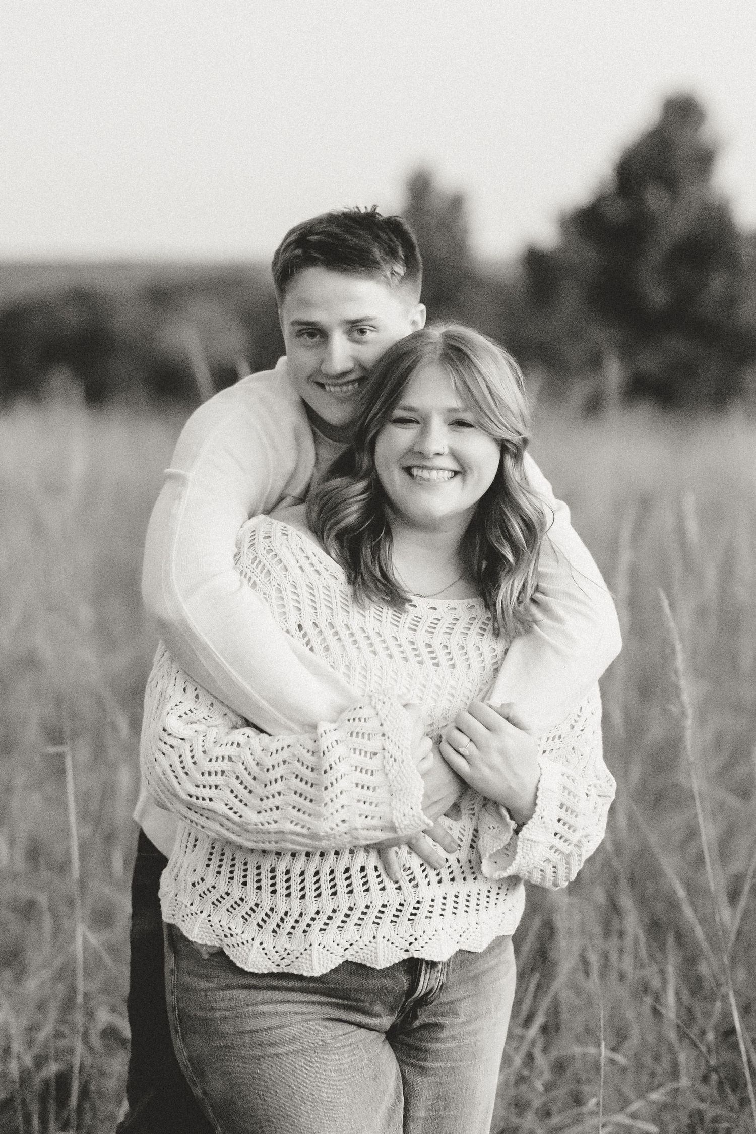 A loving couple embraces while wearing casual clothing in a field during sunset.