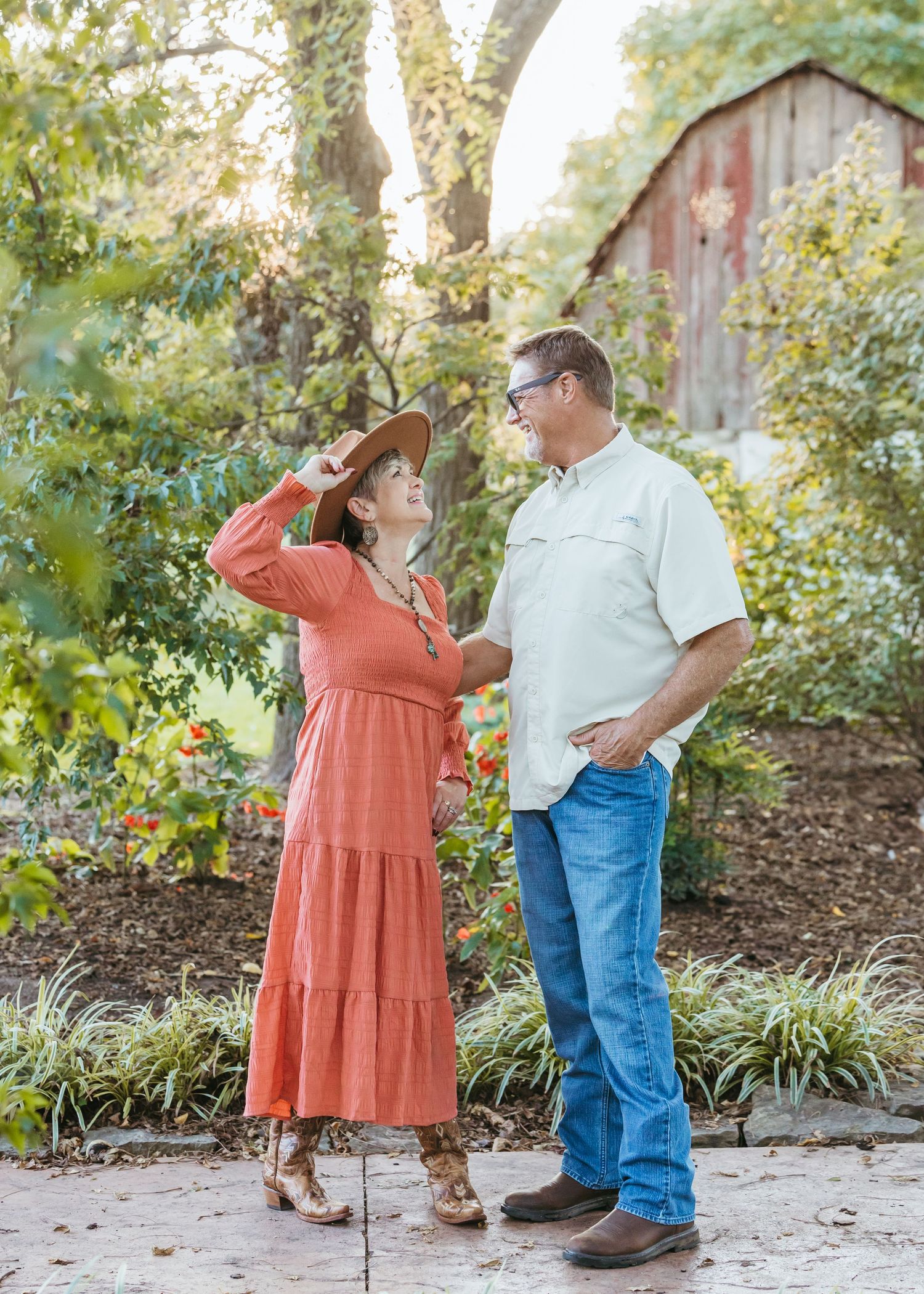 Couple in casual attire sharing playful moment in garden setting with rustic barn background.