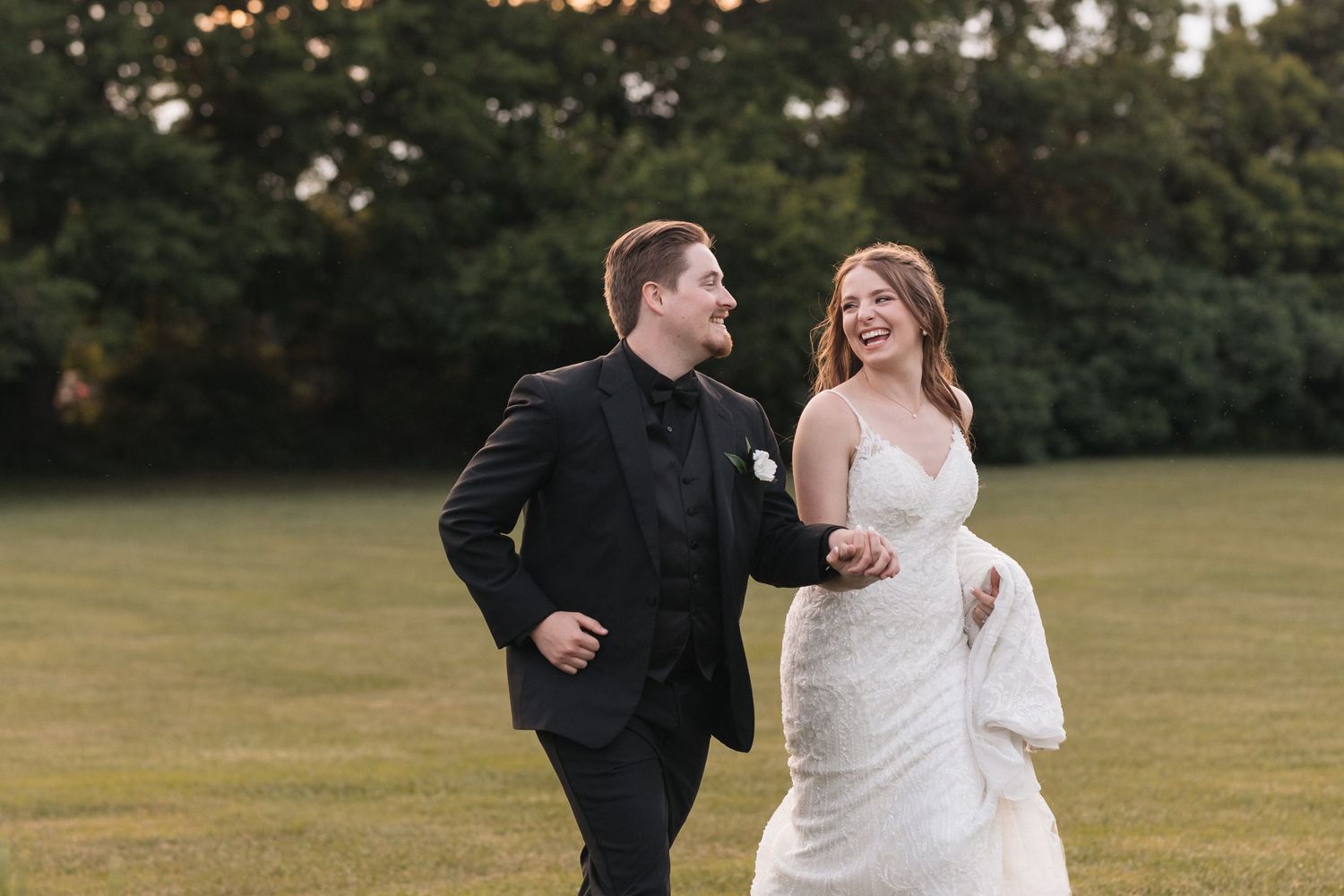 A couple runs playfully across a green field in their wedding attire.