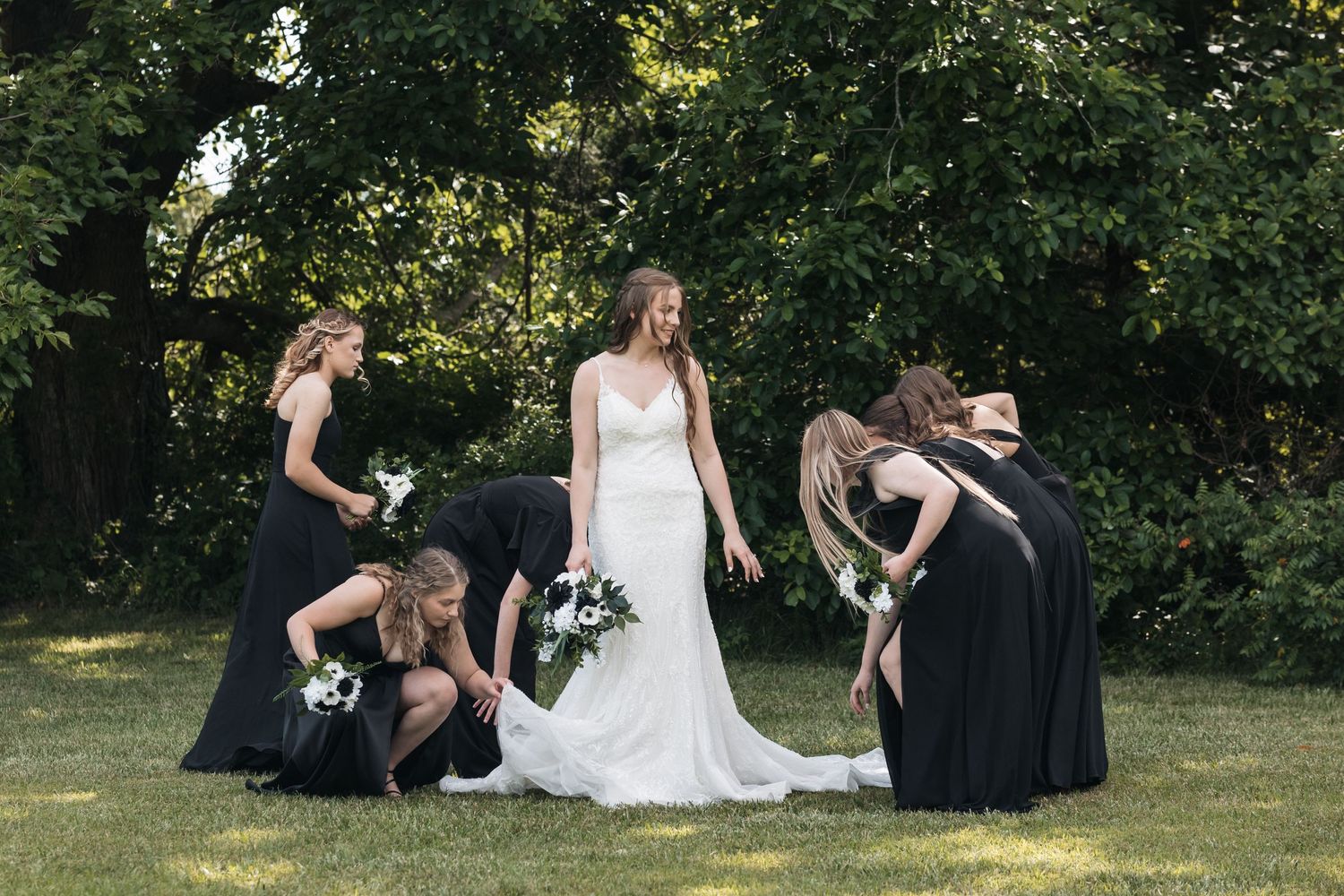 Bridesmaids in black dresses help adjust the bride's gown during an outdoor photo session.