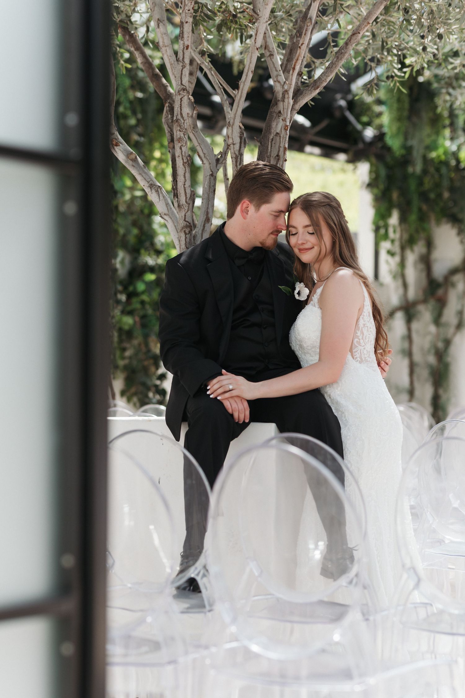 Artistic wedding photo through window frame showing couple in tender embrace among ghost chairs.