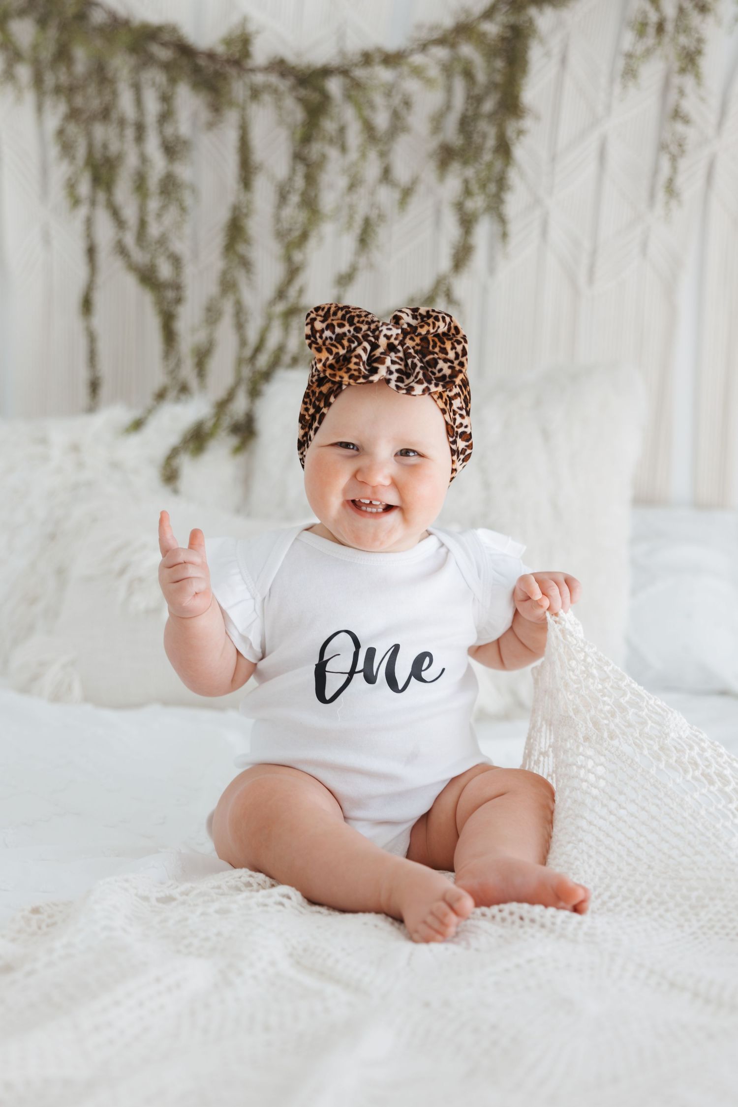 Baby wearing leopard print headband and 'One' onesie sitting on white bedding.