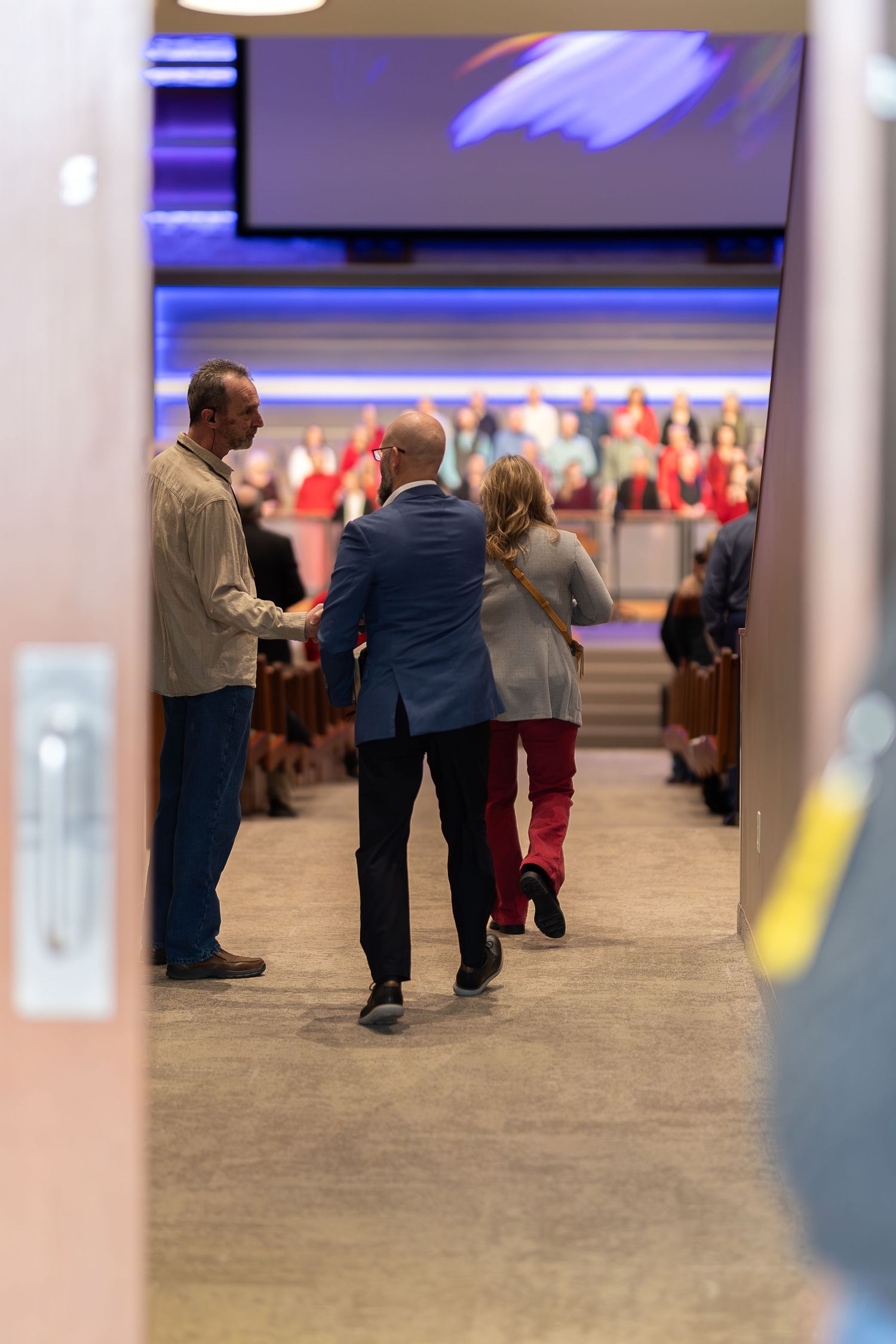 People gather in church hallway viewed through doorway with stage visible in background.