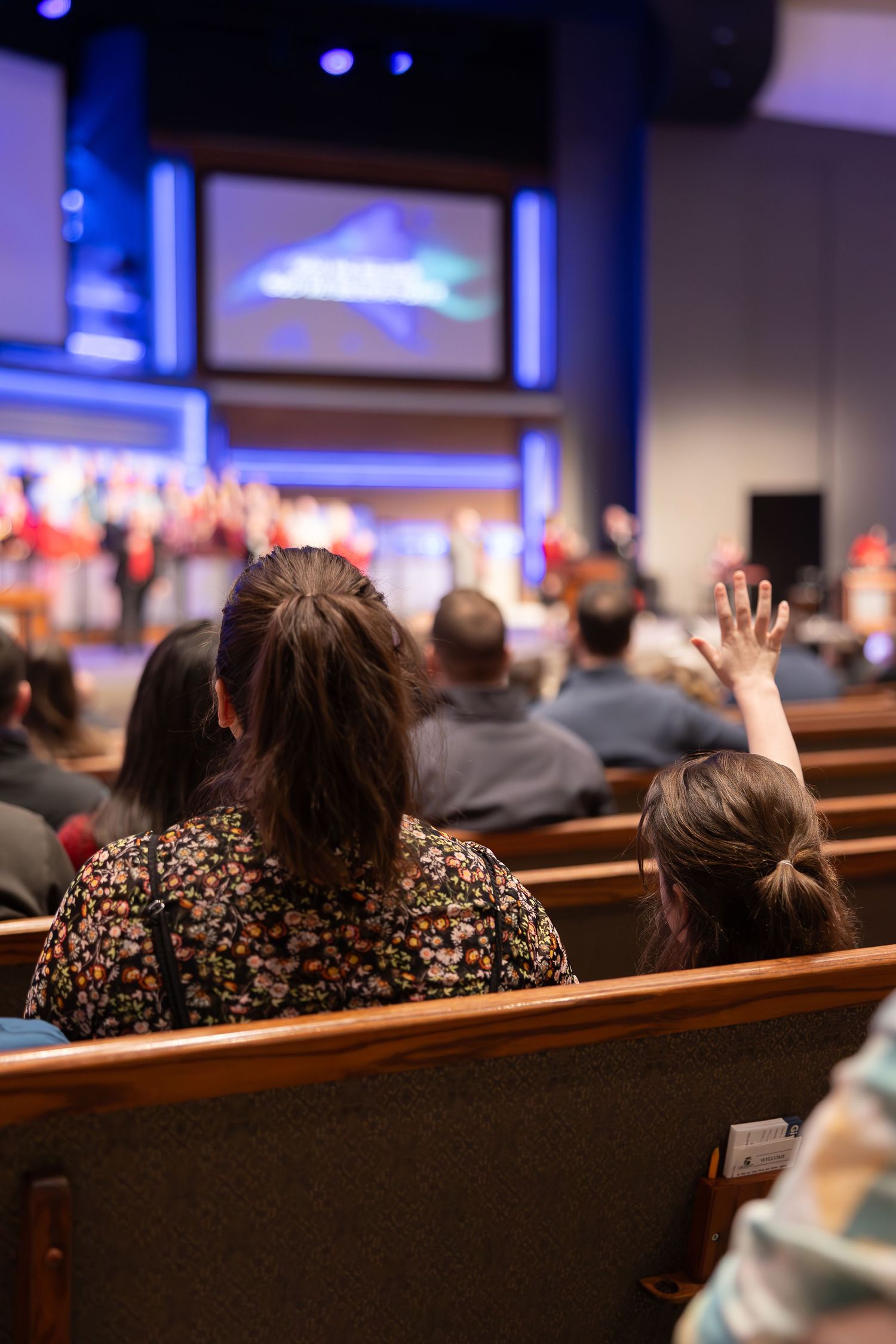 Worshippers raise hands during church service with video screens visible in background.
