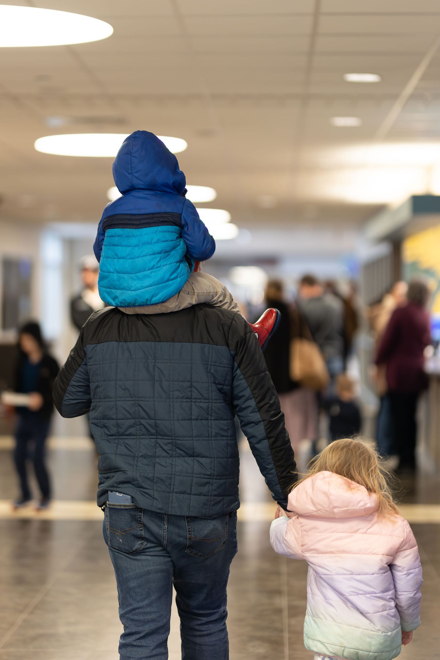 Parent carrying child on shoulders while another child walks beside them in indoor venue.