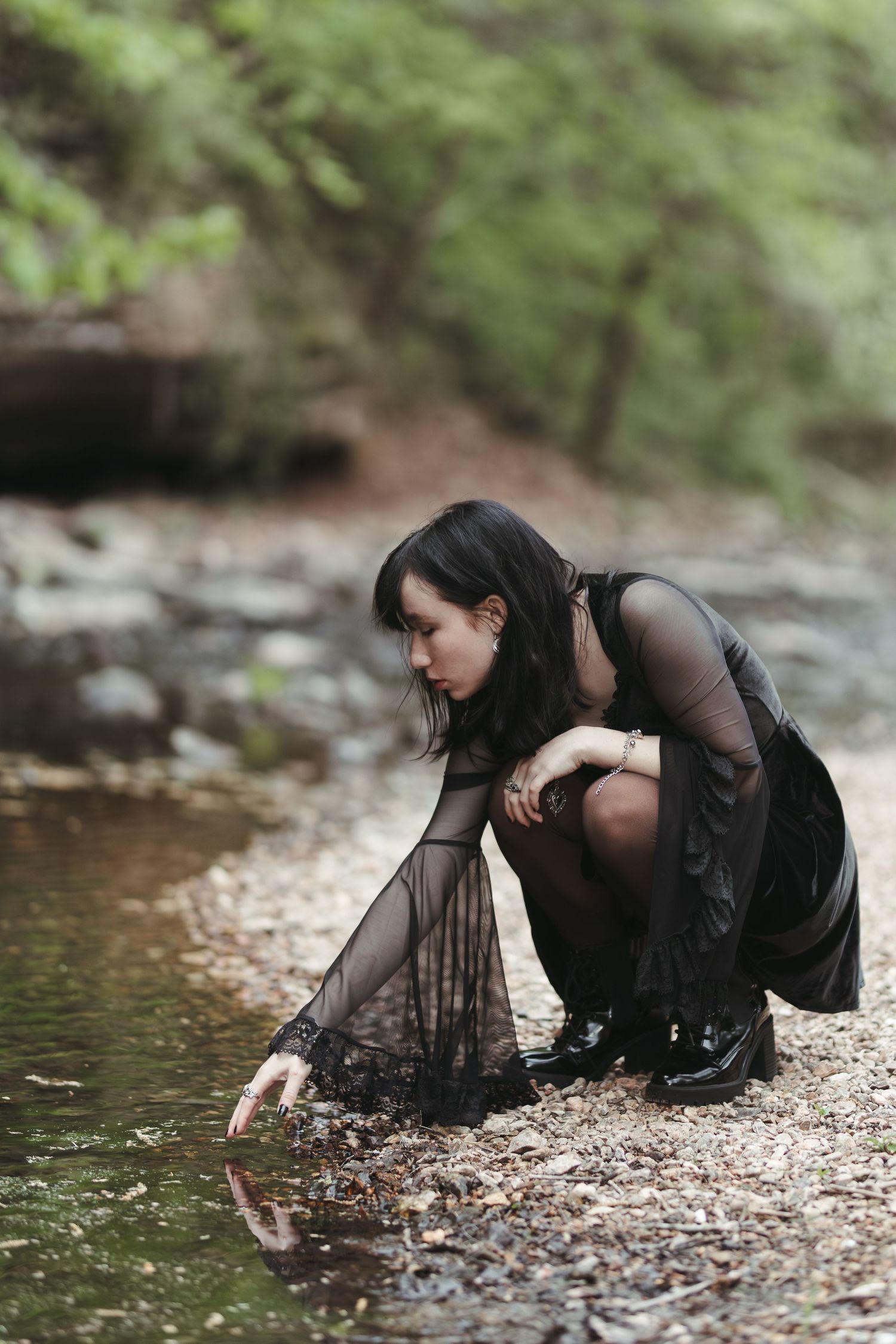 A person in dark clothing crouches by a stream reaching toward the water in a forest setting.