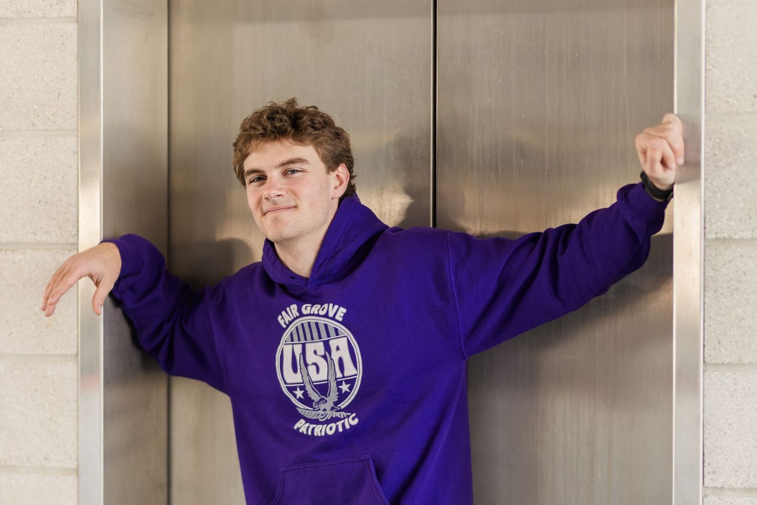 Casual pose against elevator wearing purple university sweatshirt with athletic logo.