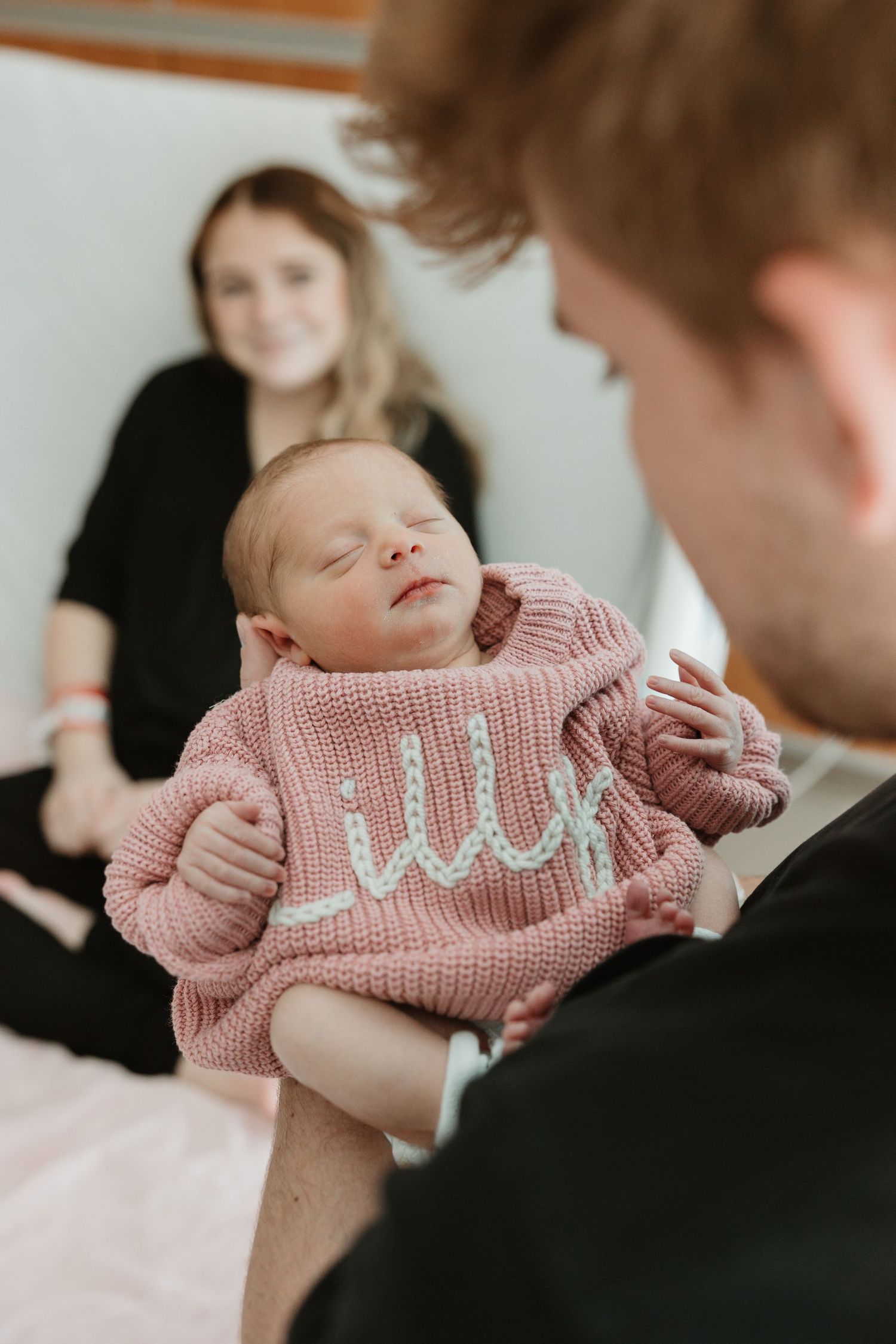 Sleeping newborn baby wearing pink knit sweater held by parent with another adult in background.