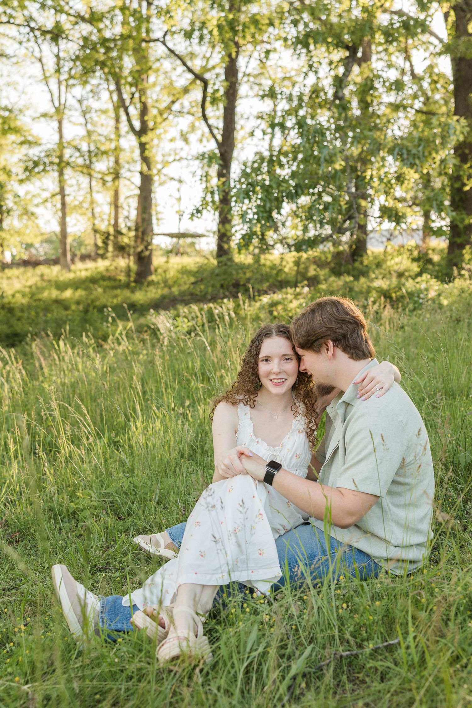 A romantic couple sits together on grassy hill during golden hour with birch trees in background.