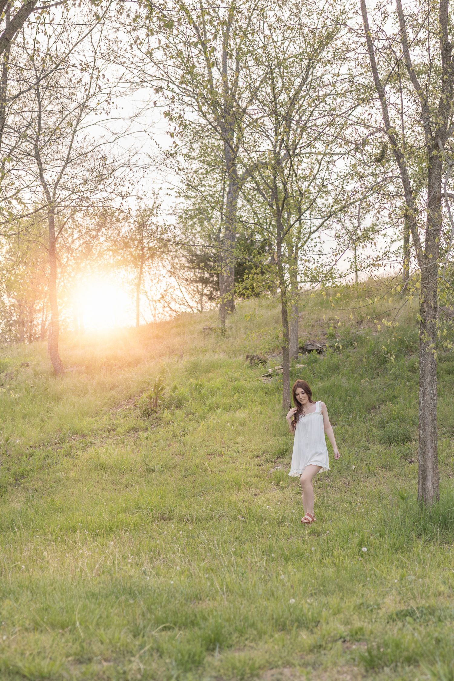 A person in a white dress walks down a grassy hillside during golden hour with trees and sunlight in the background.