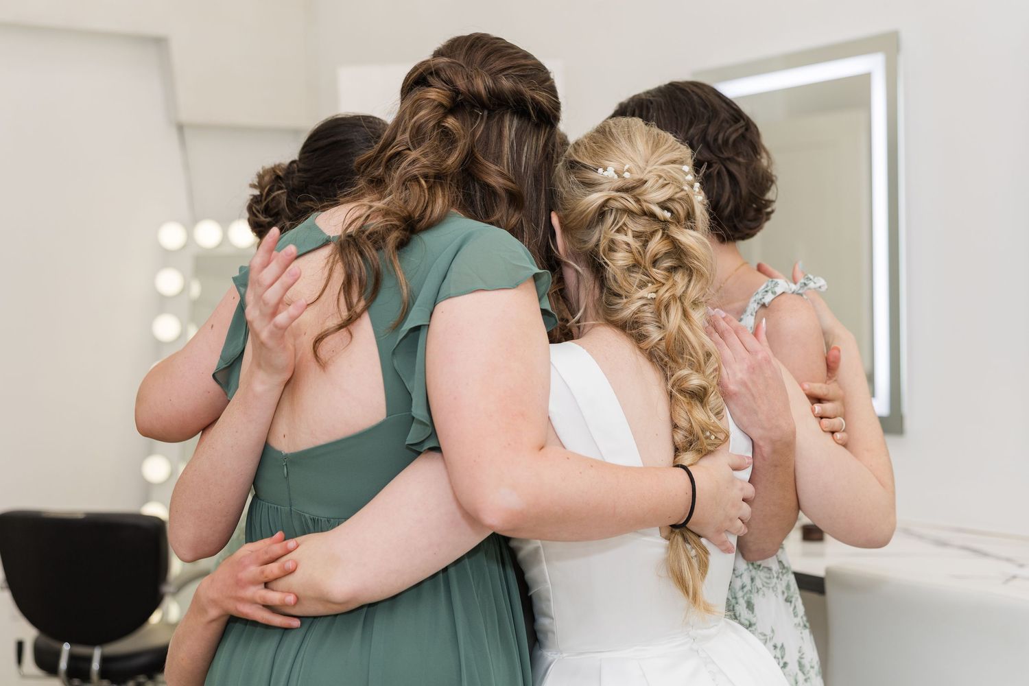 Bride and bridesmaids in green dresses share a group hug from behind in a bridal suite with mirror.