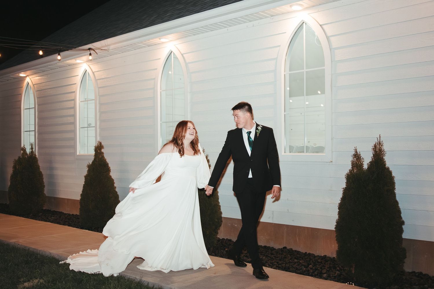 Bride and groom holding hands walking outside a white chapel at night, bride in flowing white gown.