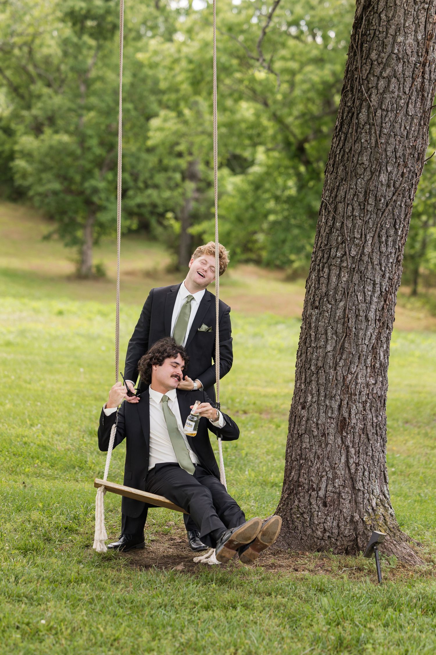 Two grooms in black suits laugh on a wooden rope swing tied to a tree in a lush green outdoor setting.