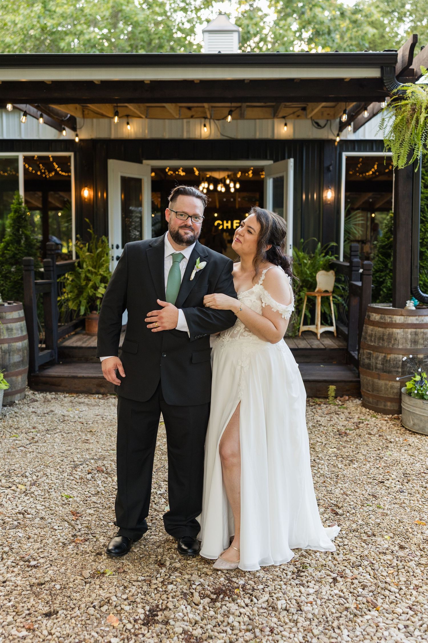 Newlyweds share a laugh outside a rustic venue decorated with string lights and wine barrels.