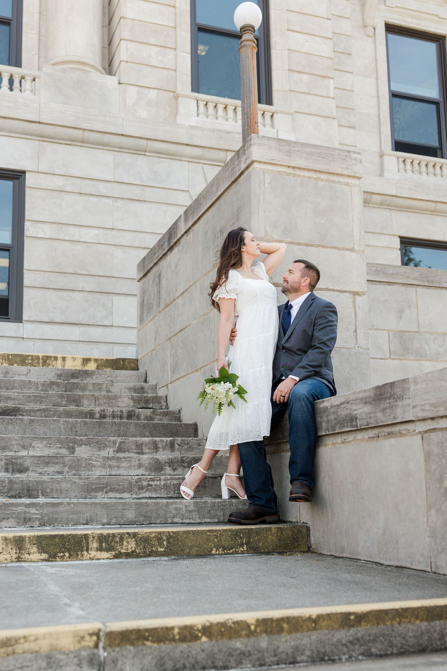 A bride and groom sit romantically together on stone steps outside a grand courthouse building during their wedding shoot.