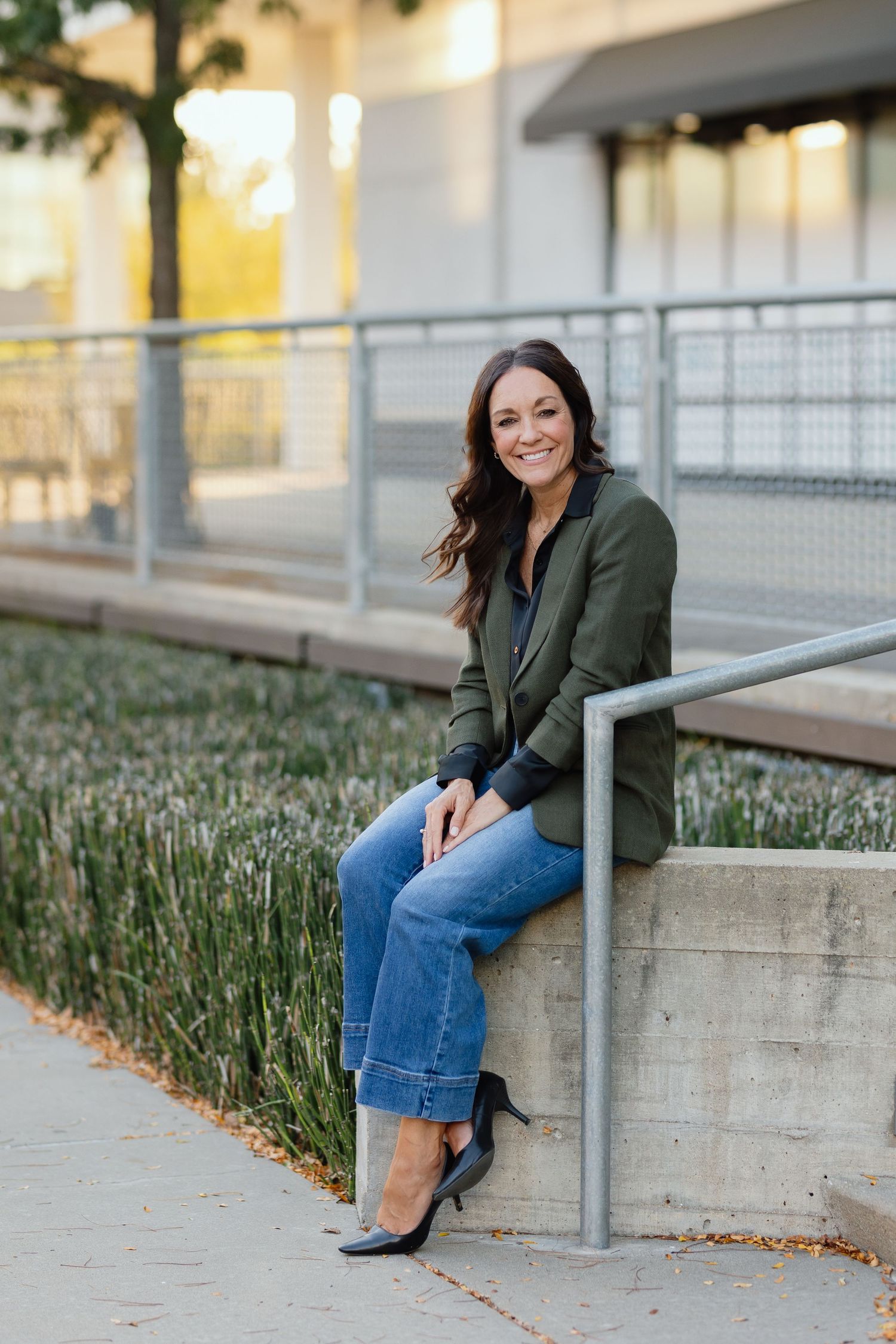 A casual autumn outfit with an olive green jacket, blue jeans and black heels on a sidewalk with plants.