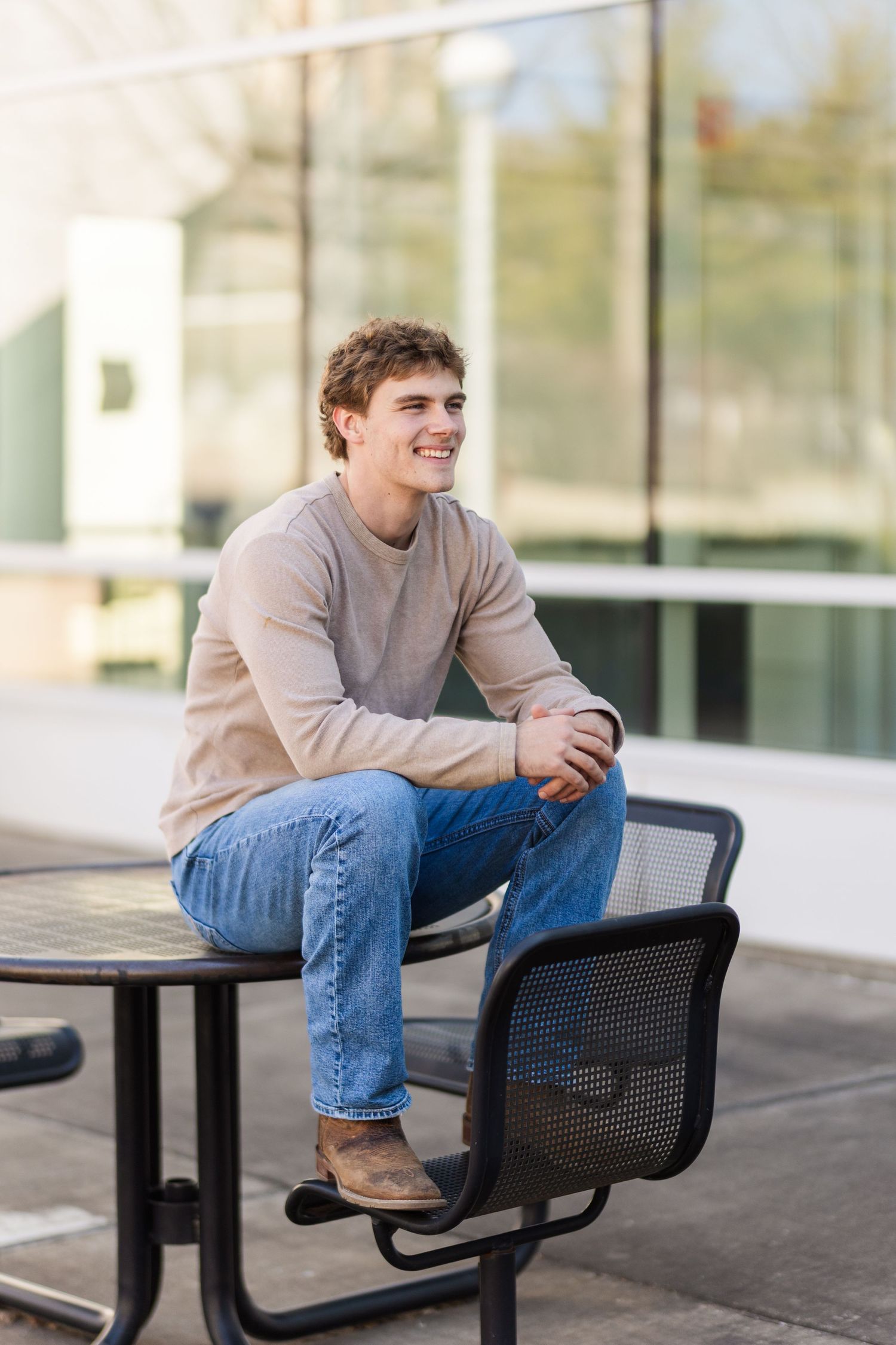A person in a beige sweater and jeans sits casually on an outdoor bench at what appears to be a campus or office setting.