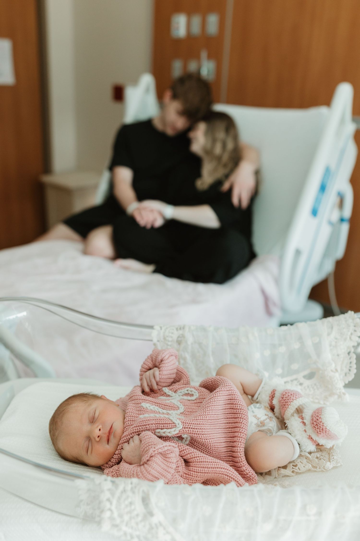 A sleeping newborn baby wearing a pink sweater lies on white bedding in a hospital room.