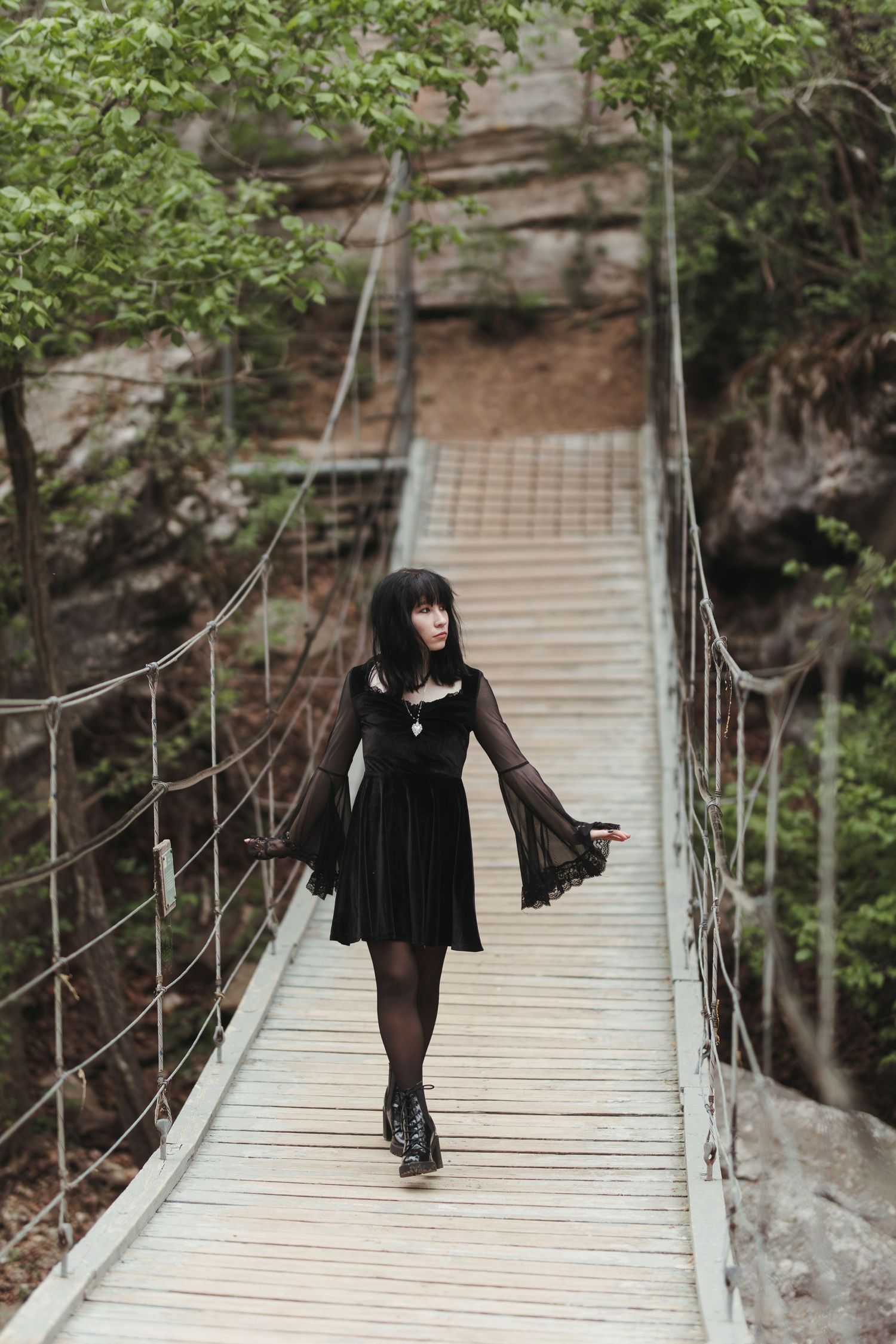 A person in a black gothic dress stands on a wooden suspension bridge surrounded by lush greenery.