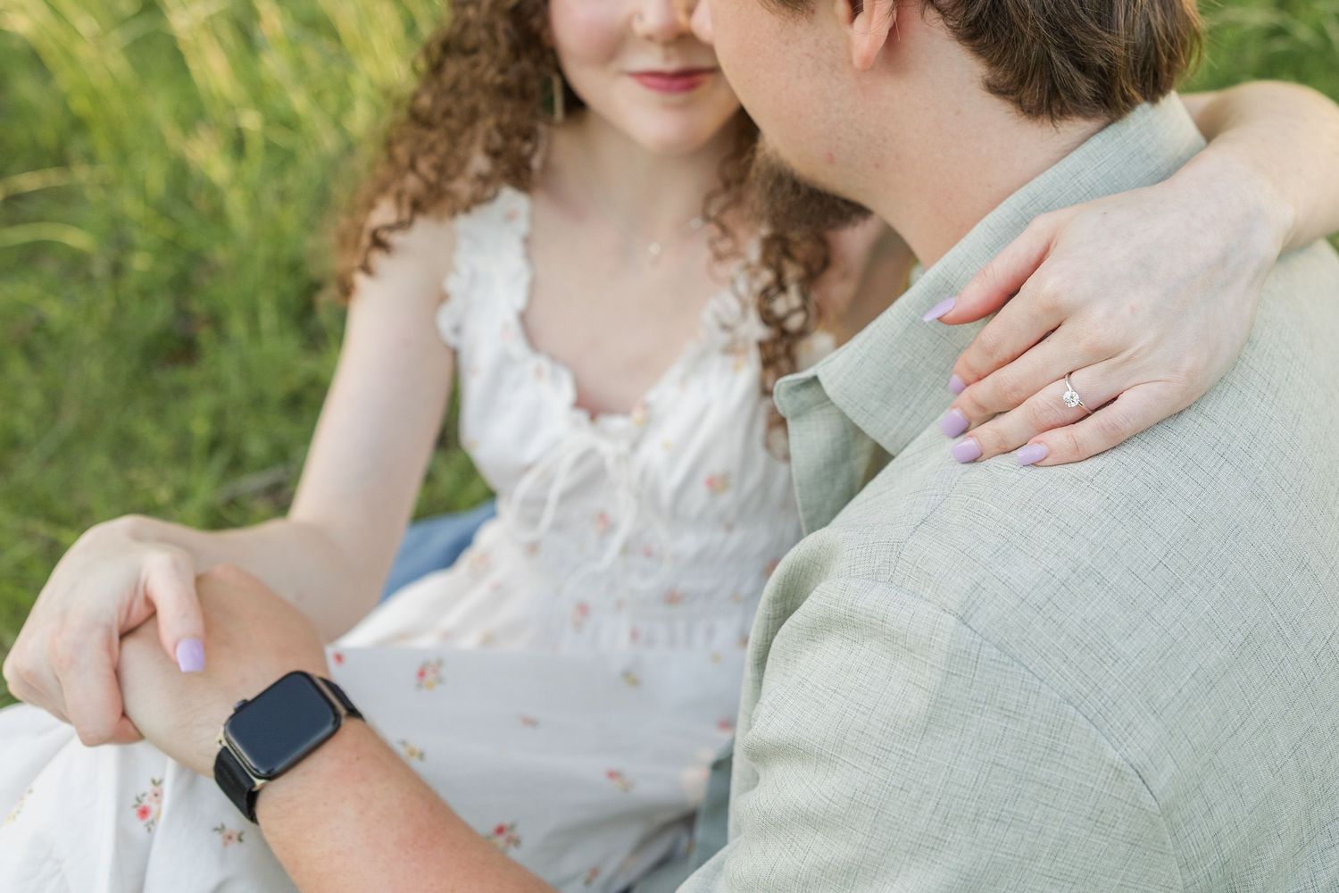 Couple sitting in grass outdoors, wearing white dress and gray shirt, with hands touching affectionately.