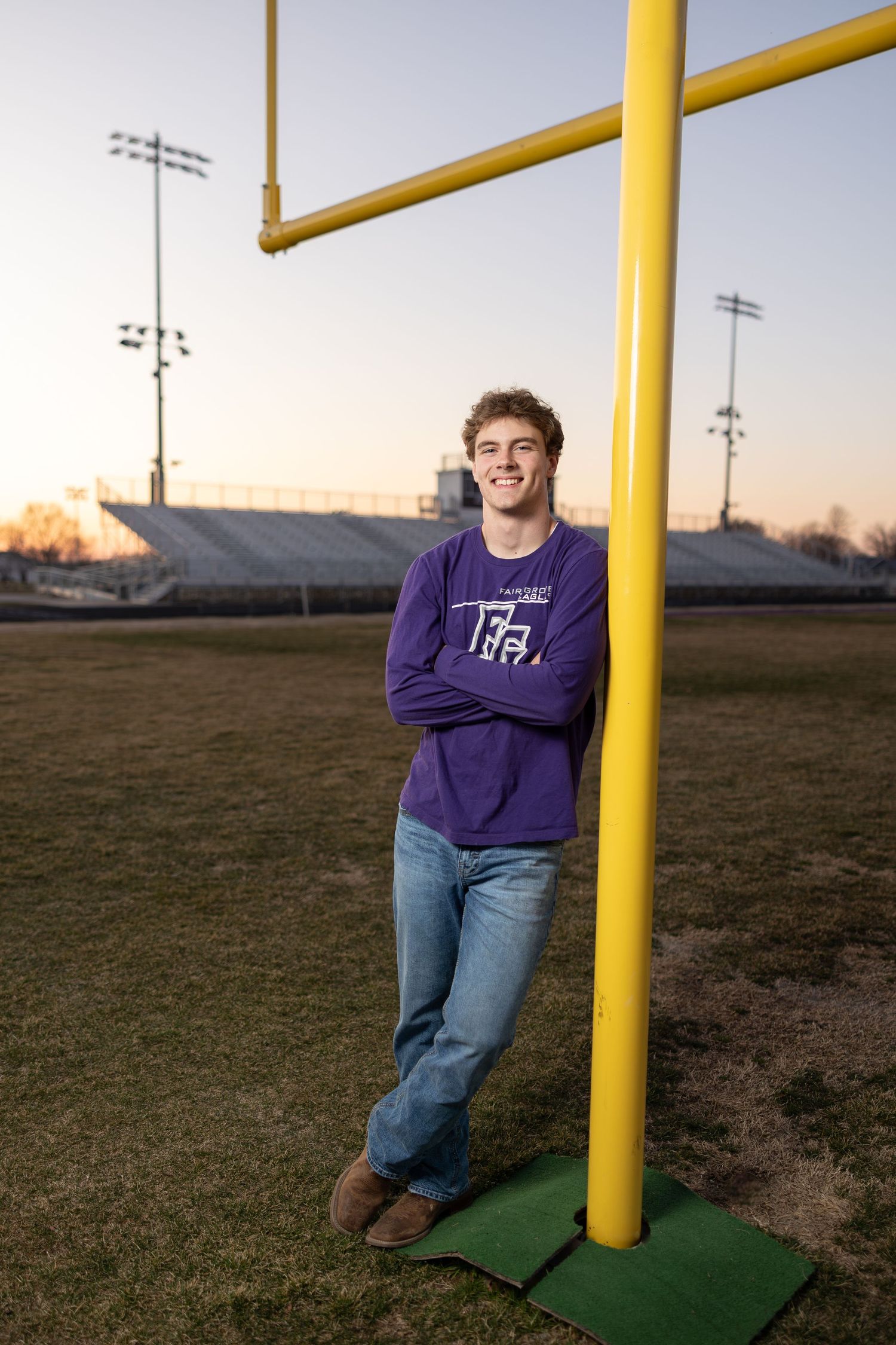 Person in purple hoodie leaning against yellow football goalpost on athletic field at sunset.