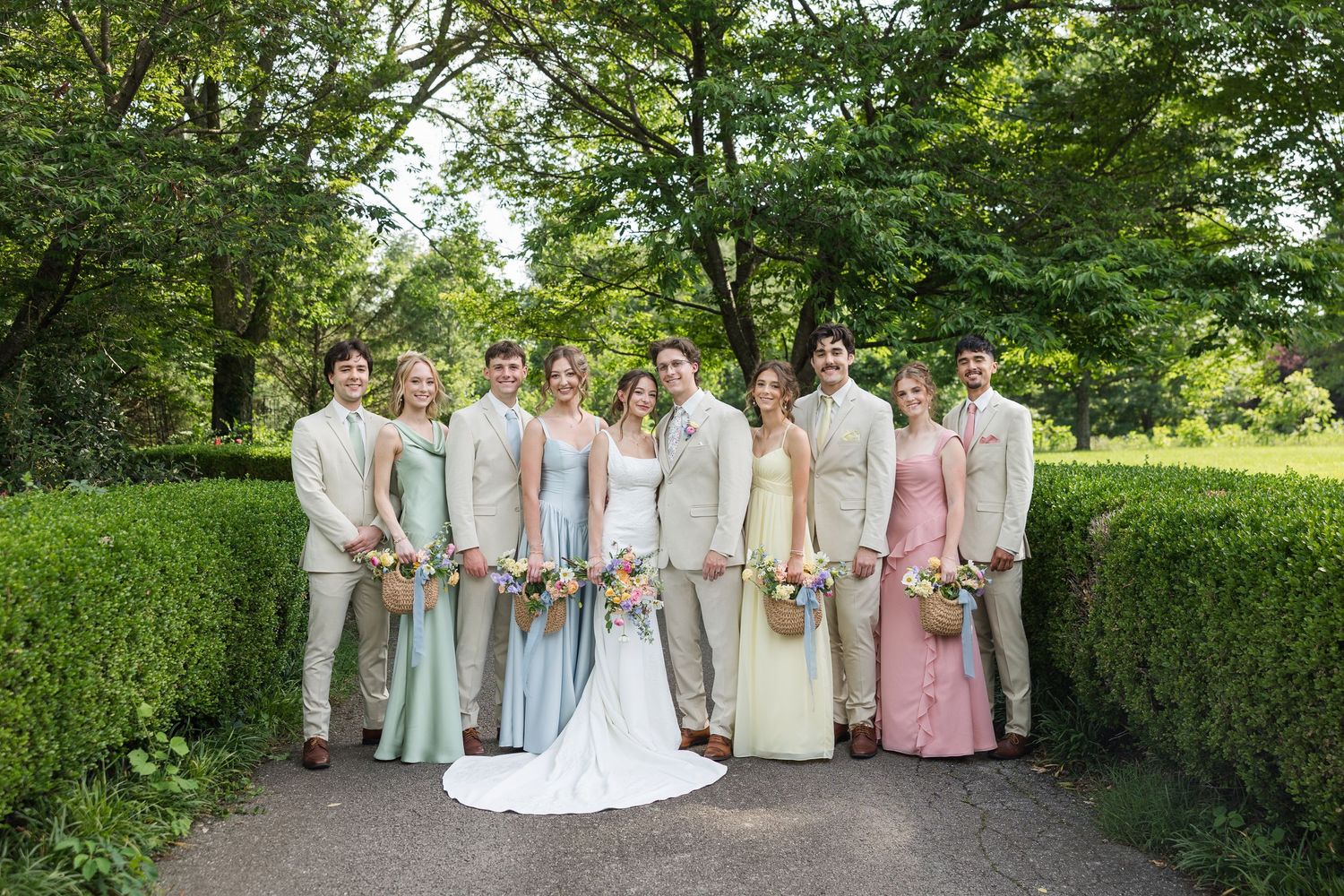 Wedding party posing outdoors in garden, bridesmaids in pastel gowns and groomsmen in tan suits flanking bride and groom.