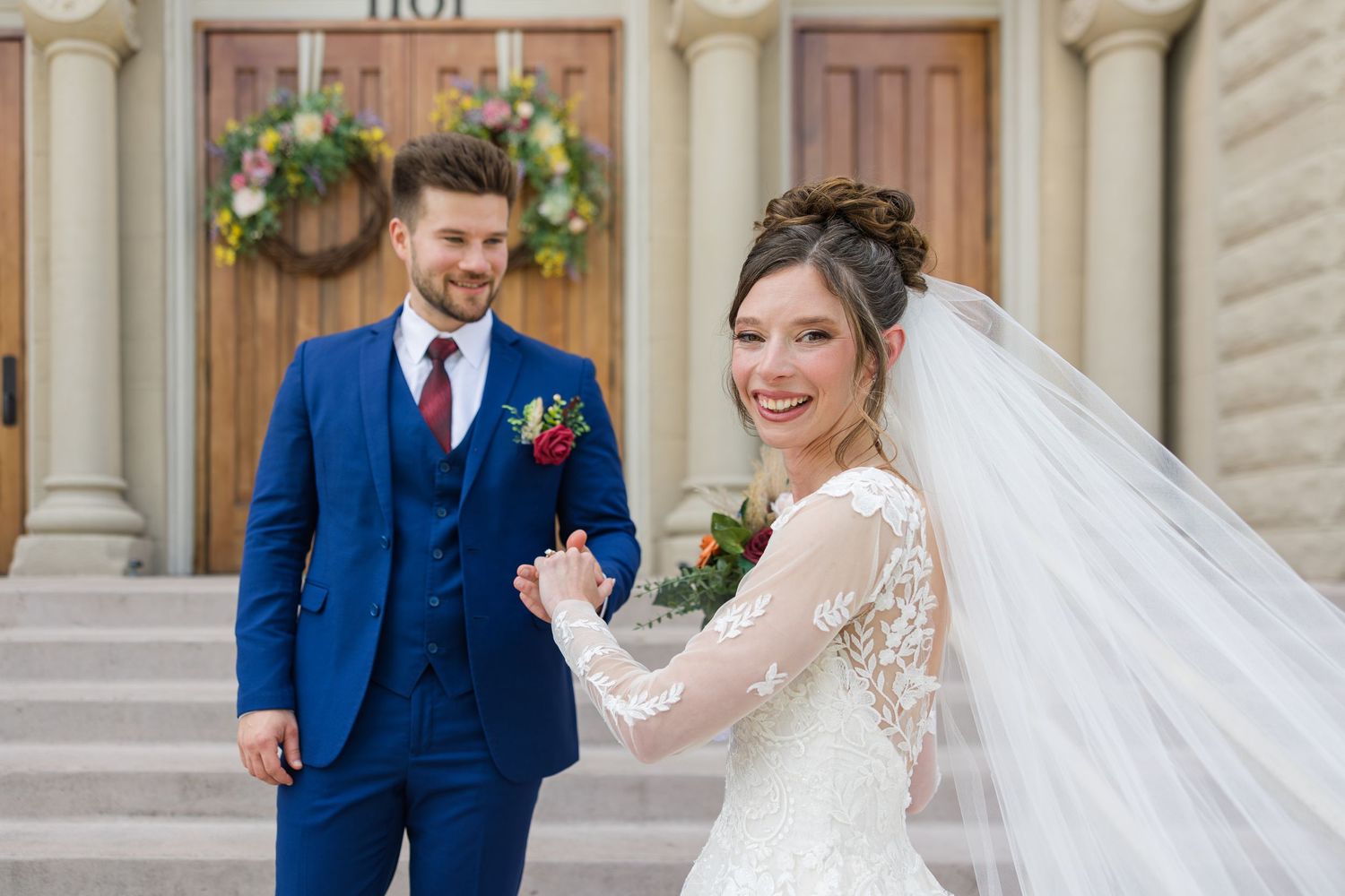 Smiling bride in lace gown with veil holds grooms hand in blue suit on church steps.