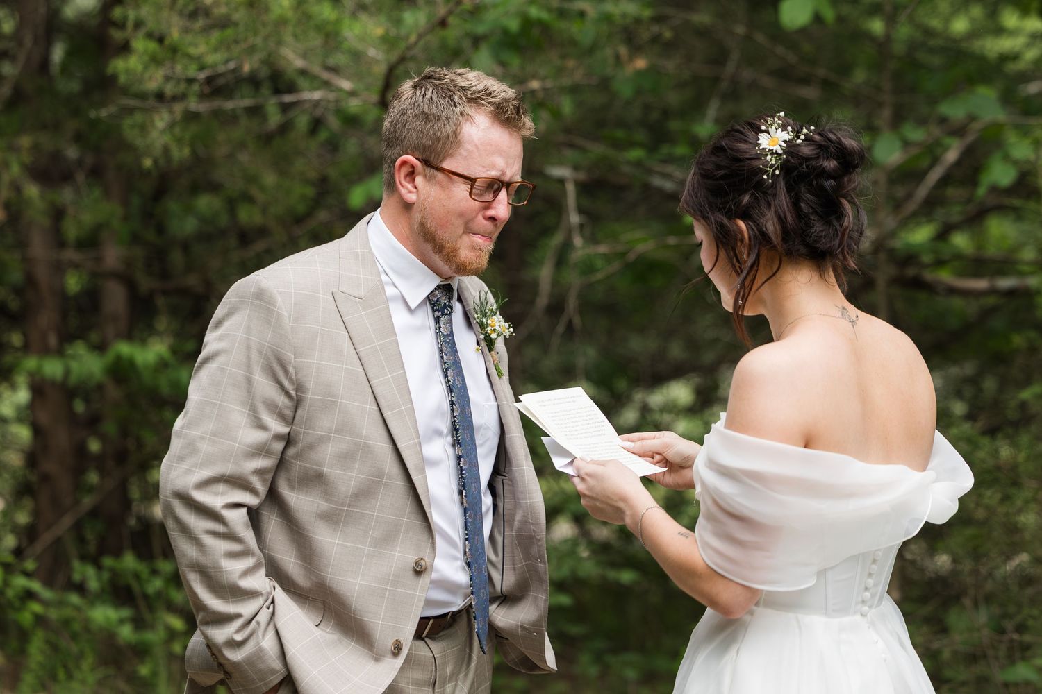 Bride reads wedding vows to groom in tan suit outdoors at woodland ceremony.